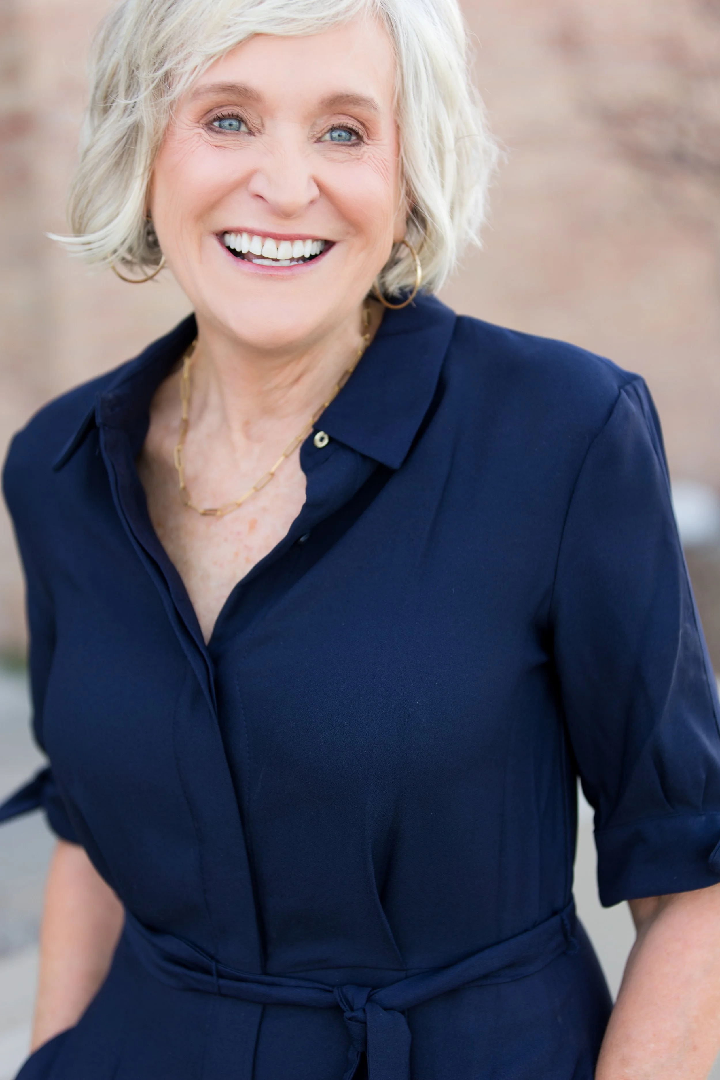 Close-up of a smiling woman with short blonde hair, wearing a navy blue dress with a tied waist, gold jewelry, and hoop earrings, standing outdoors with a blurred background.