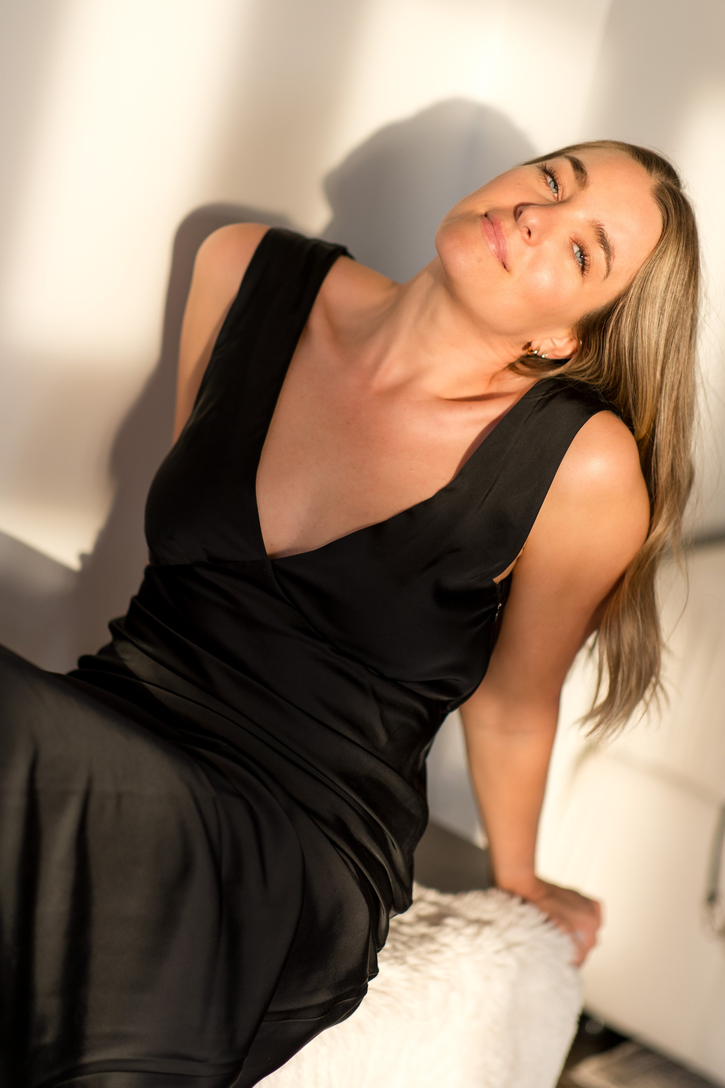 A woman in a black sleeveless dress sitting on a bed, smiling softly, with her head tilted to the side and long blonde hair.