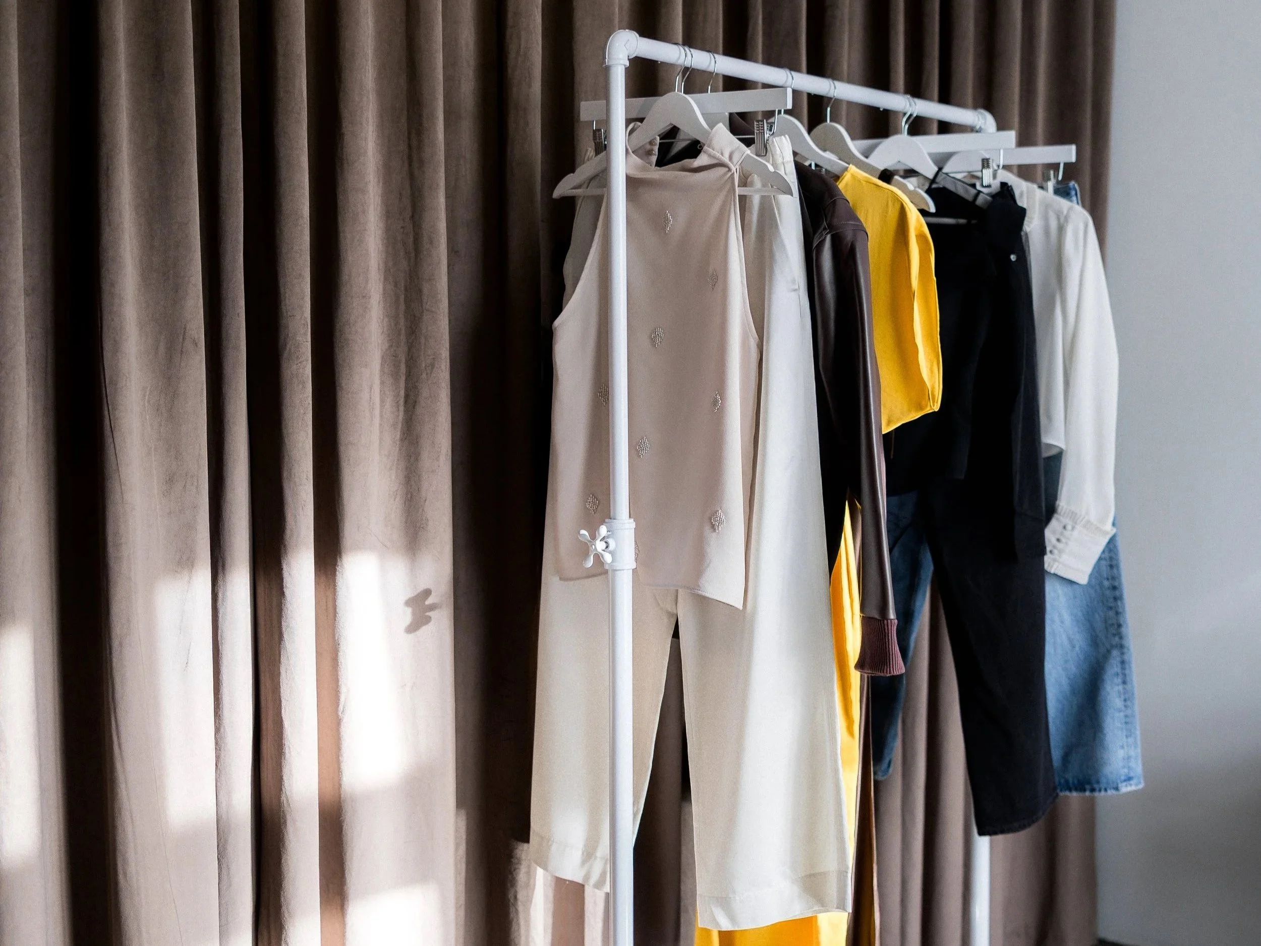 Clothes hanging on a white clothing rack in front of brown curtains. Items include a sleeveless beige top, yellow pants, a dark brown jacket, a black blazer, a white shirt, and blue denim jeans.