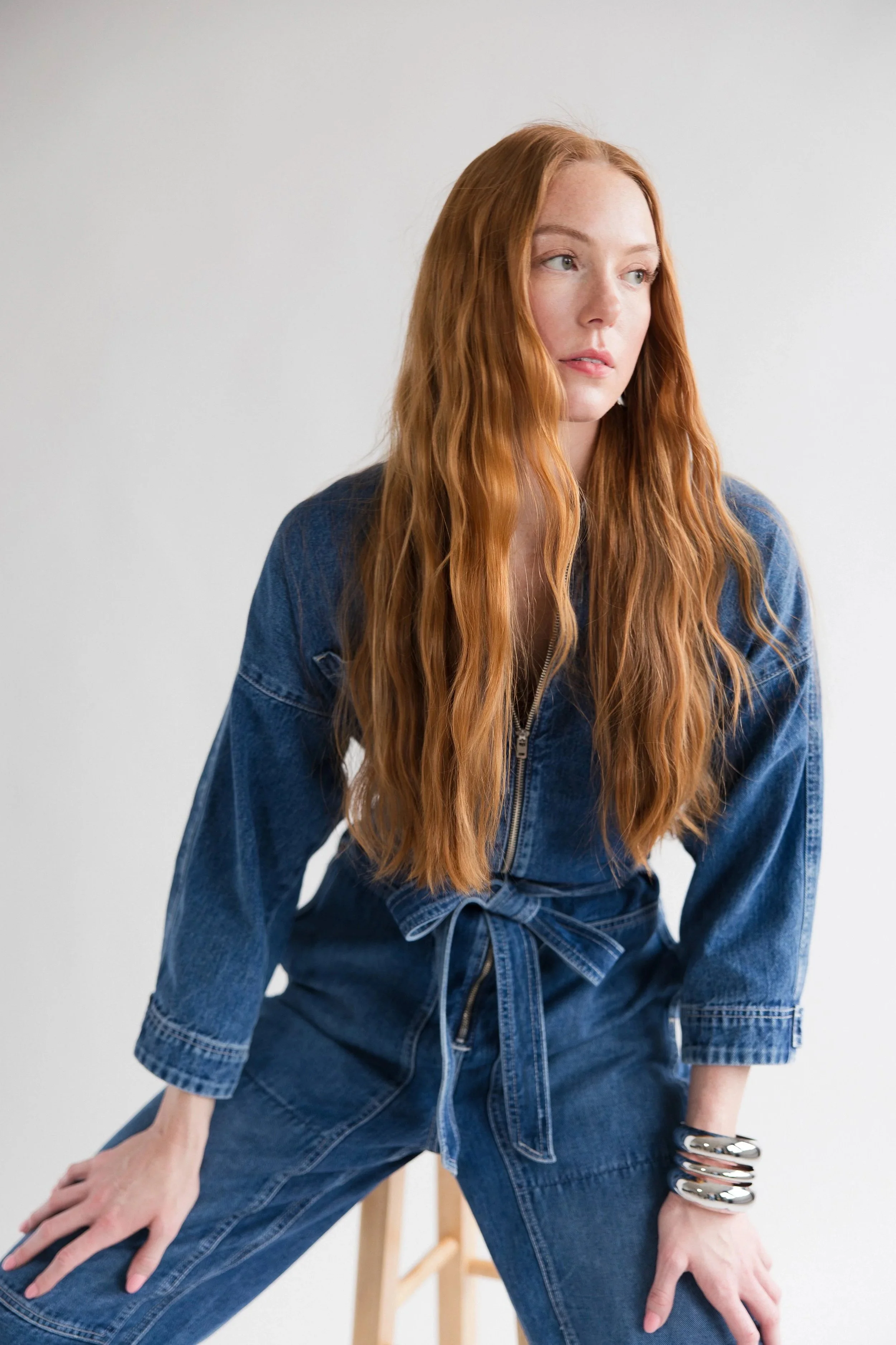 A woman with long red hair in a blue denim outfit is sitting on a wooden stool against a plain white wall. She is wearing multiple silver bangles on her right wrist.