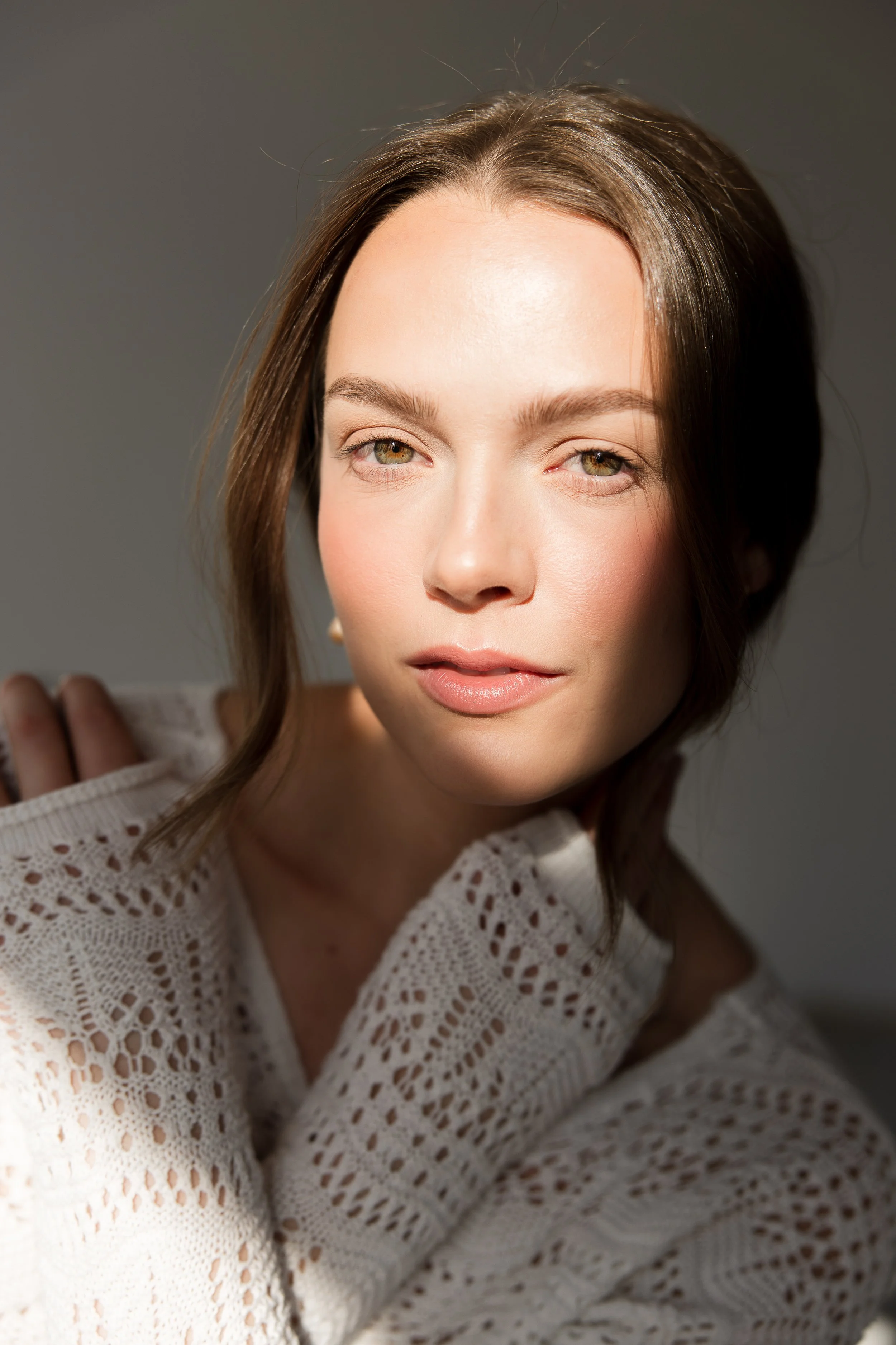 A young woman with light brown hair styled in loose waves, wearing a white knitted sweater with eyelet details, looking directly at the camera with a neutral expression.