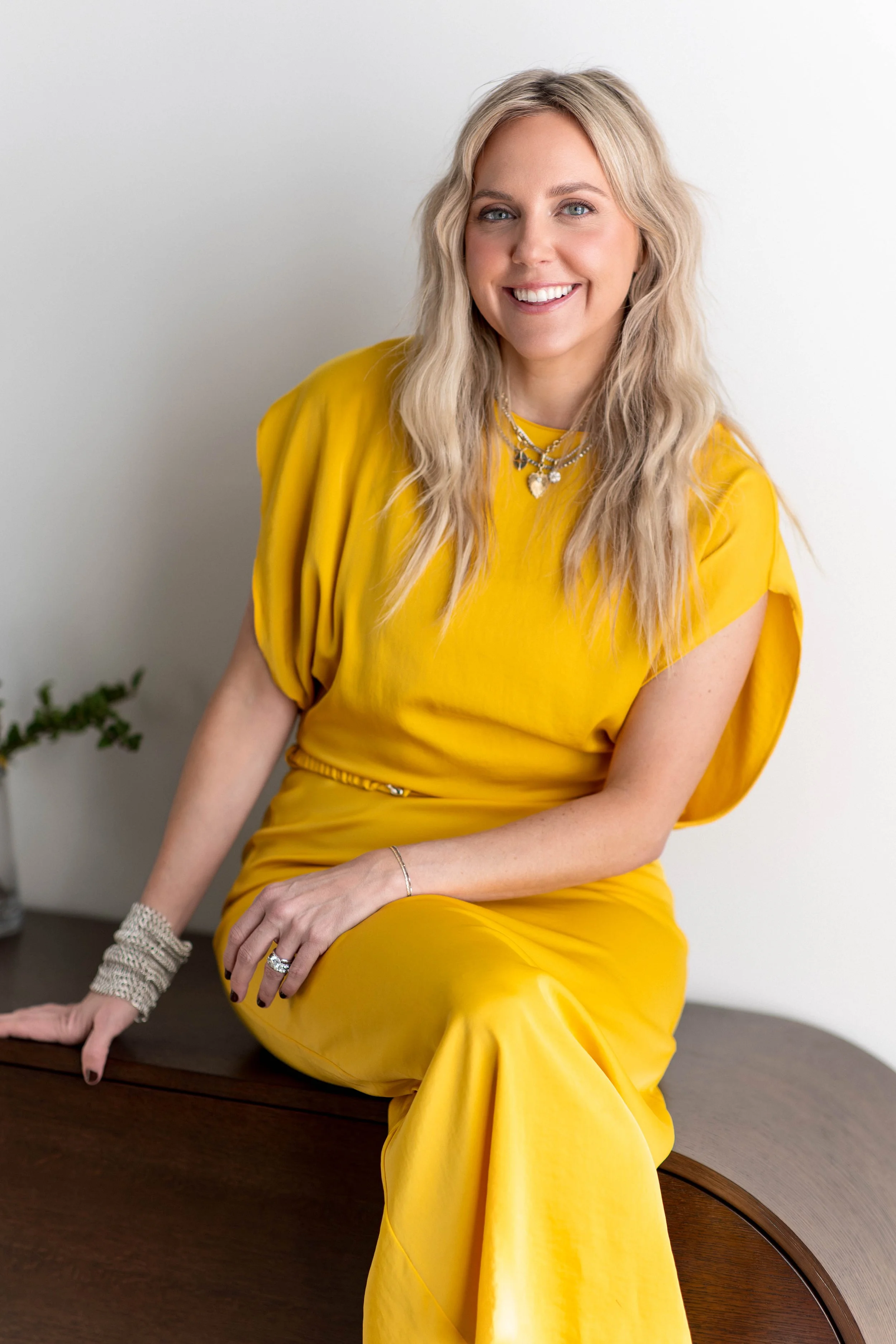 A smiling woman with blonde, wavy hair wearing a bright yellow dress and layered necklaces, sitting on a wooden table against a plain white wall.