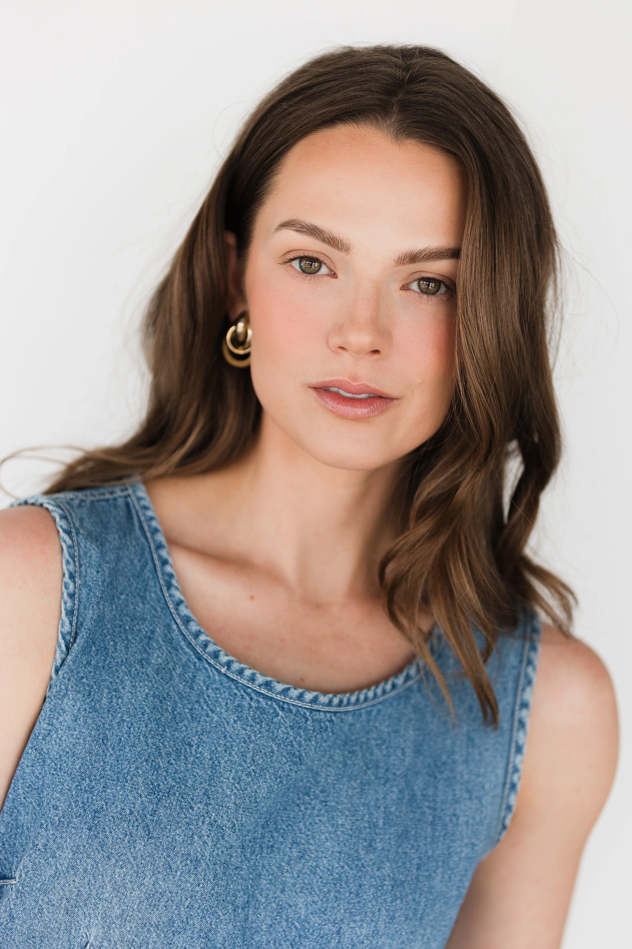 Close-up of a woman with shoulder-length brown hair, wearing gold hoop earrings and a sleeveless blue denim top, smiling softly against a plain white background.