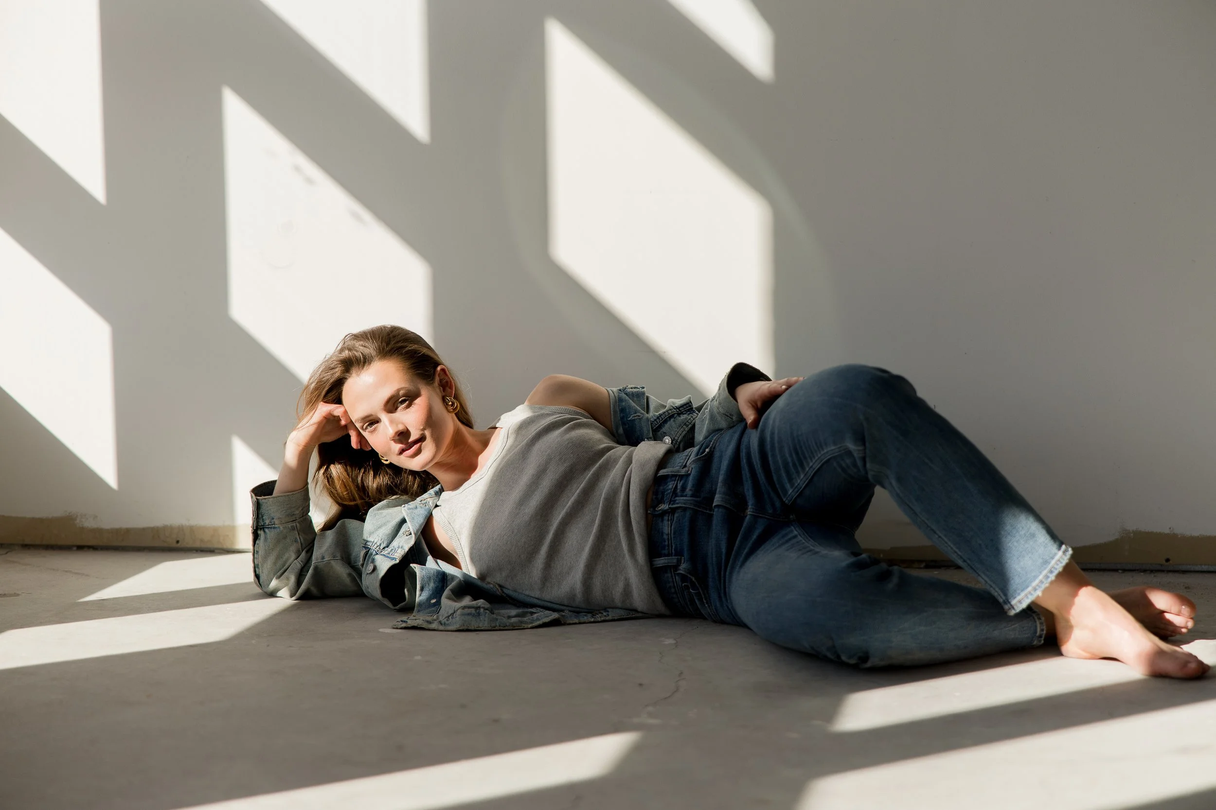 Woman lying on the floor in a brightly lit room with shadows on the wall, wearing a gray t-shirt, denim jacket, and jeans.
