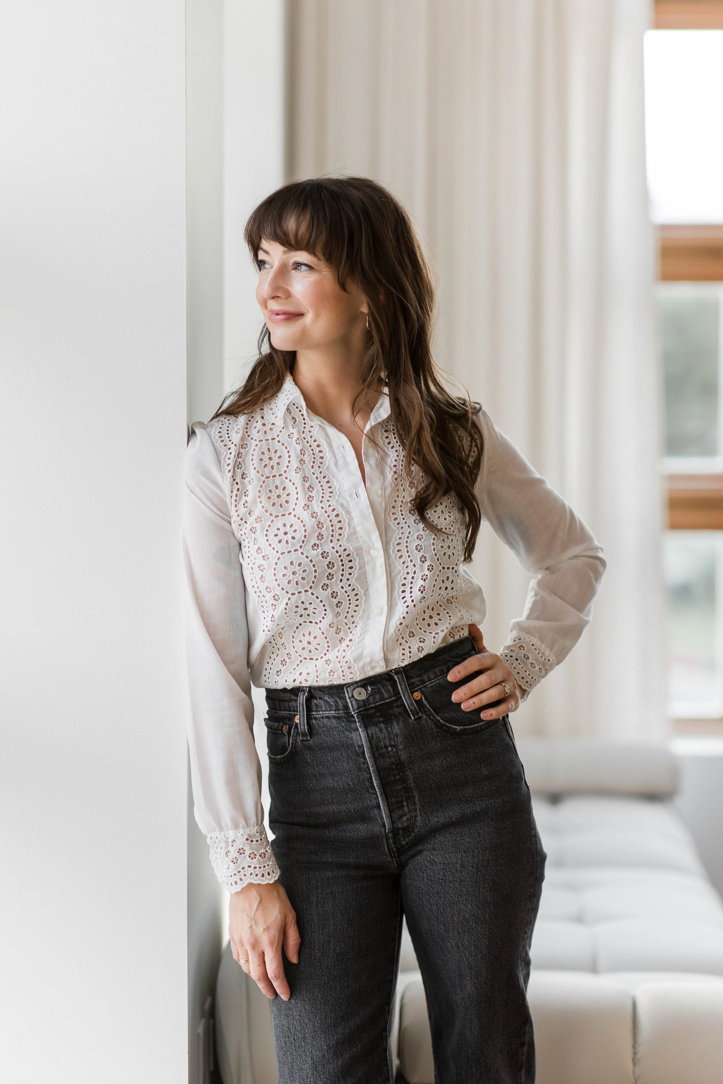 Woman with brown hair wearing a white lace blouse and black jeans standing by a white wall and window with curtains.