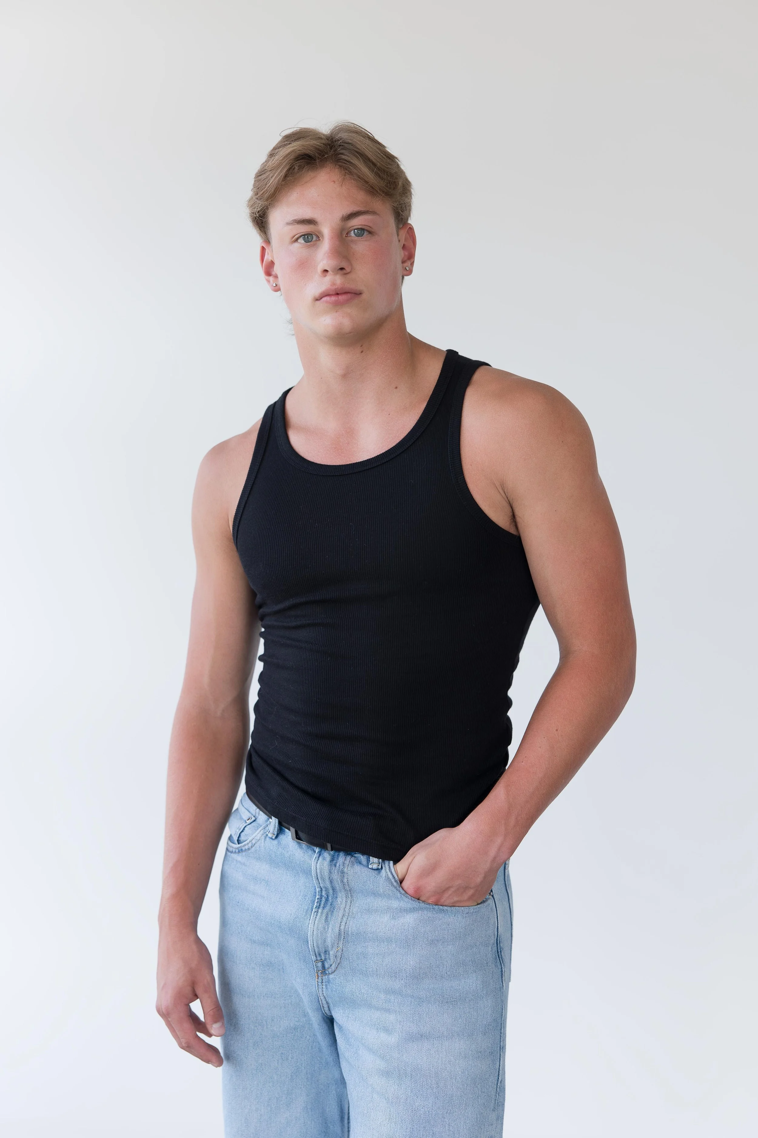 Young man with blond hair wearing a black tank top and light blue jeans, standing against a plain white background.