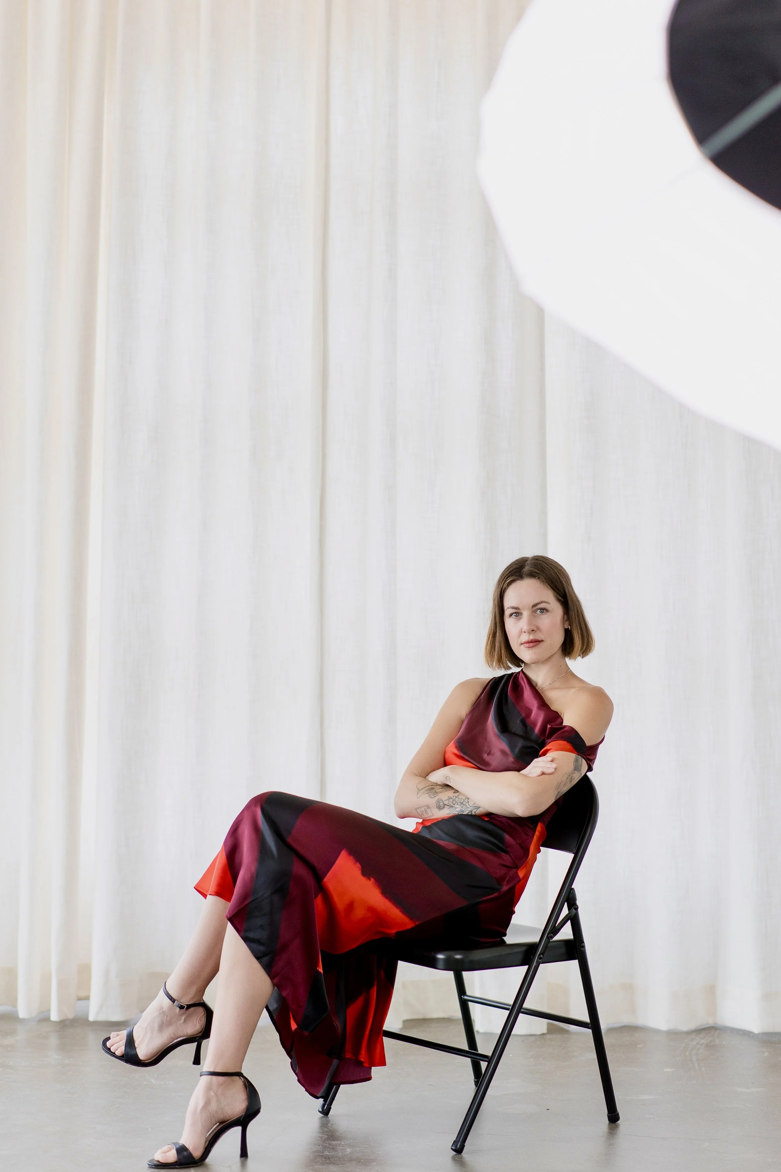 A woman with shoulder-length brown hair wearing a red and black dress and black high heels sitting with crossed arms on a black chair in front of a white curtain, with a large white photography umbrella partially visible in the upper right corner.