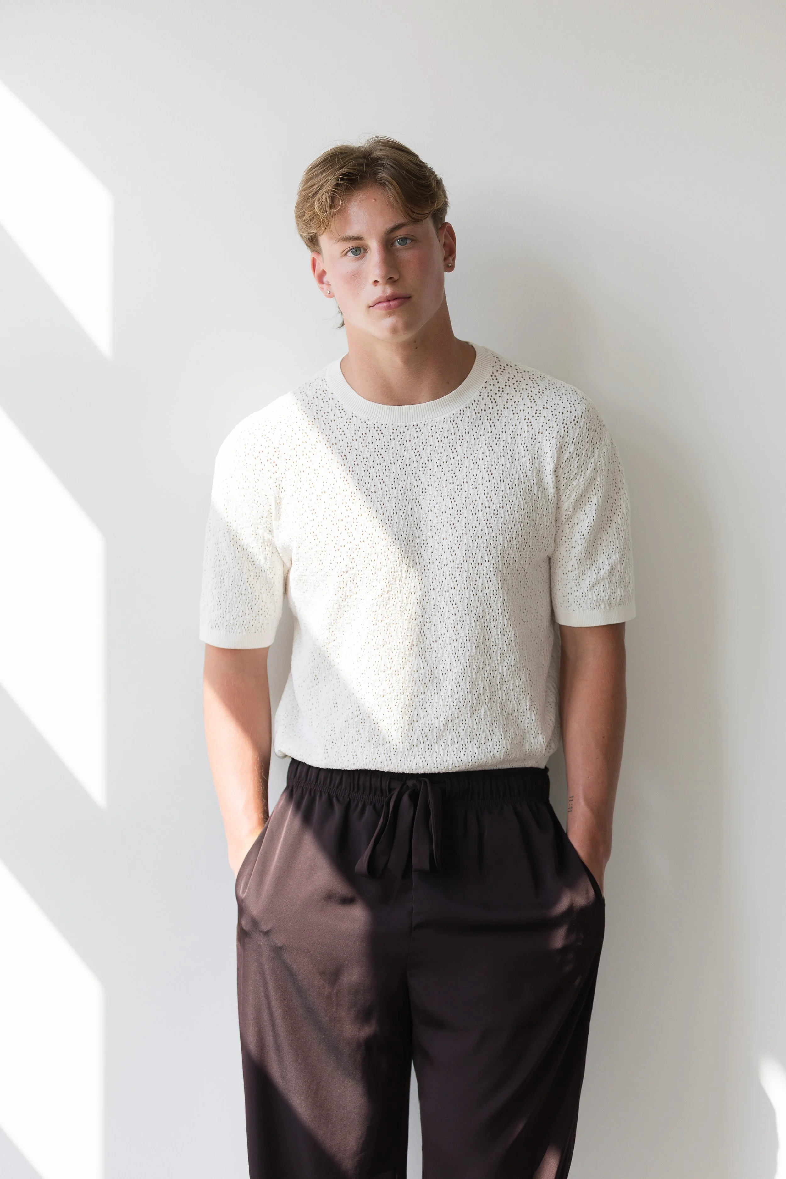 A young man with light brown hair and earrings, wearing a white perforated short-sleeve shirt and black satin pants with a drawstring, standing against a white wall with diagonal sunlight patterns.