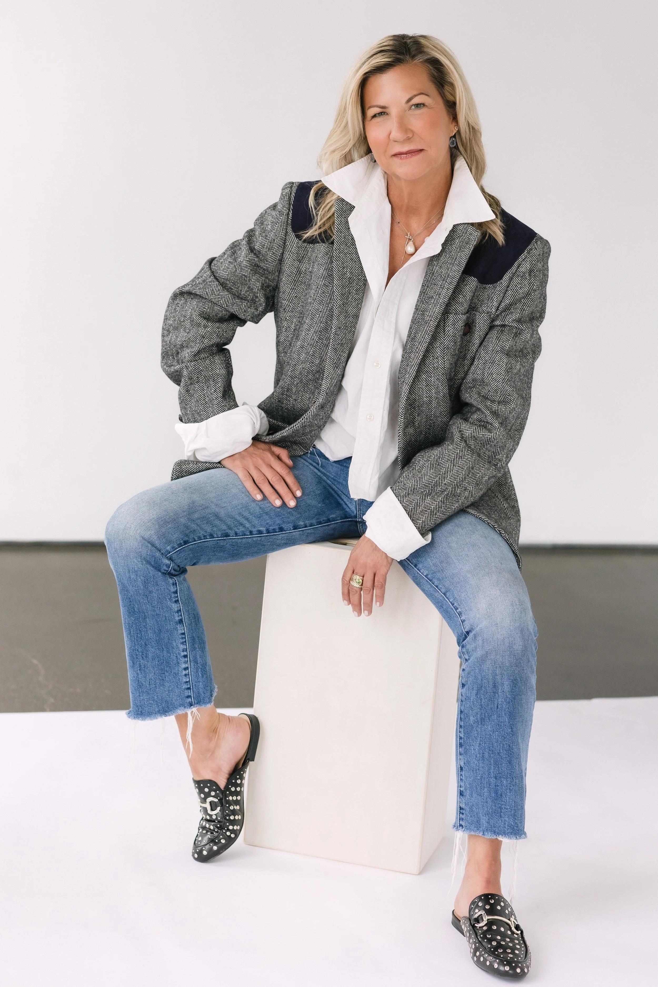 A woman wearing a blazer, white shirt, and jeans, sitting on a white block against a plain background. She is also wearing black slip-on shoes with silver studs and leopard print details. She is posing with a confident expression, with one hand on her hip and one foot resting on the block.