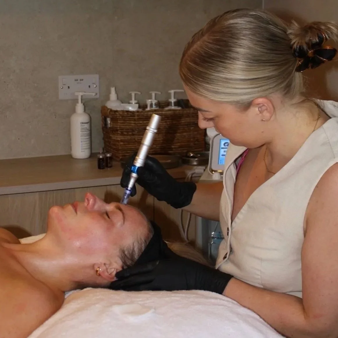 A woman receiving a cosmetic injection treatment in a clinic, lying on the table with her eyes closed, while a practitioner, wearing gloves, holds a syringe.