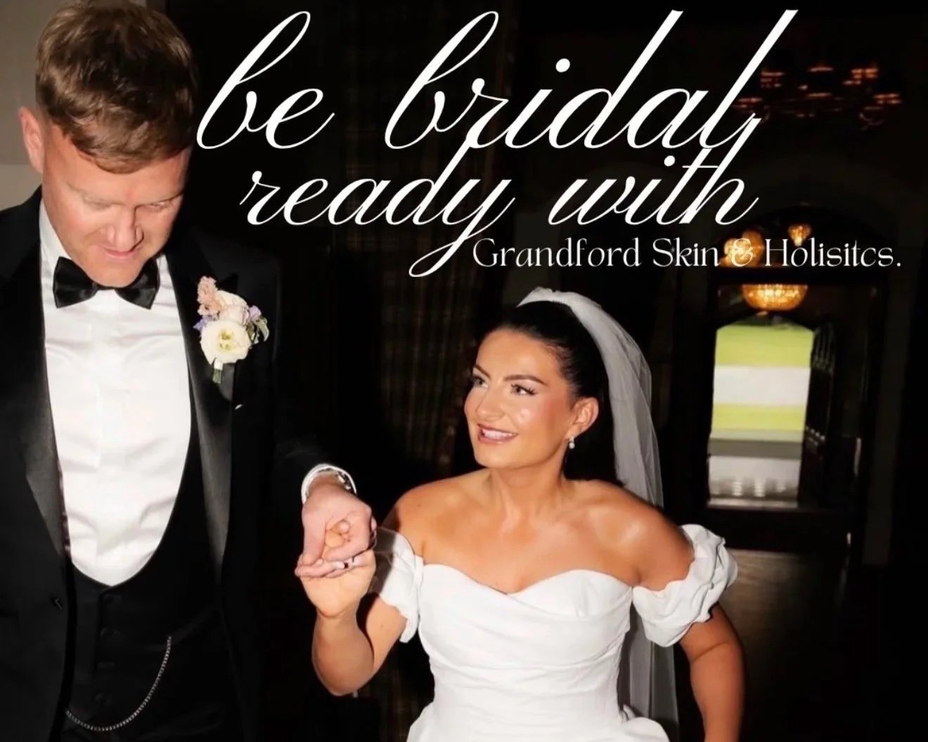 A bride and a groom holding hands, dressed in wedding attire, in a dimly lit indoor setting with a window in the background. The groom is wearing a black tuxedo with a bow tie and boutonniere, smiling down at the bride. The bride is wearing a white off-the-shoulder wedding dress, a veil, and earrings, looking up and smiling at the groom. Text overlay says, 'be bridal ready with Grandford Skin & Holistics.'