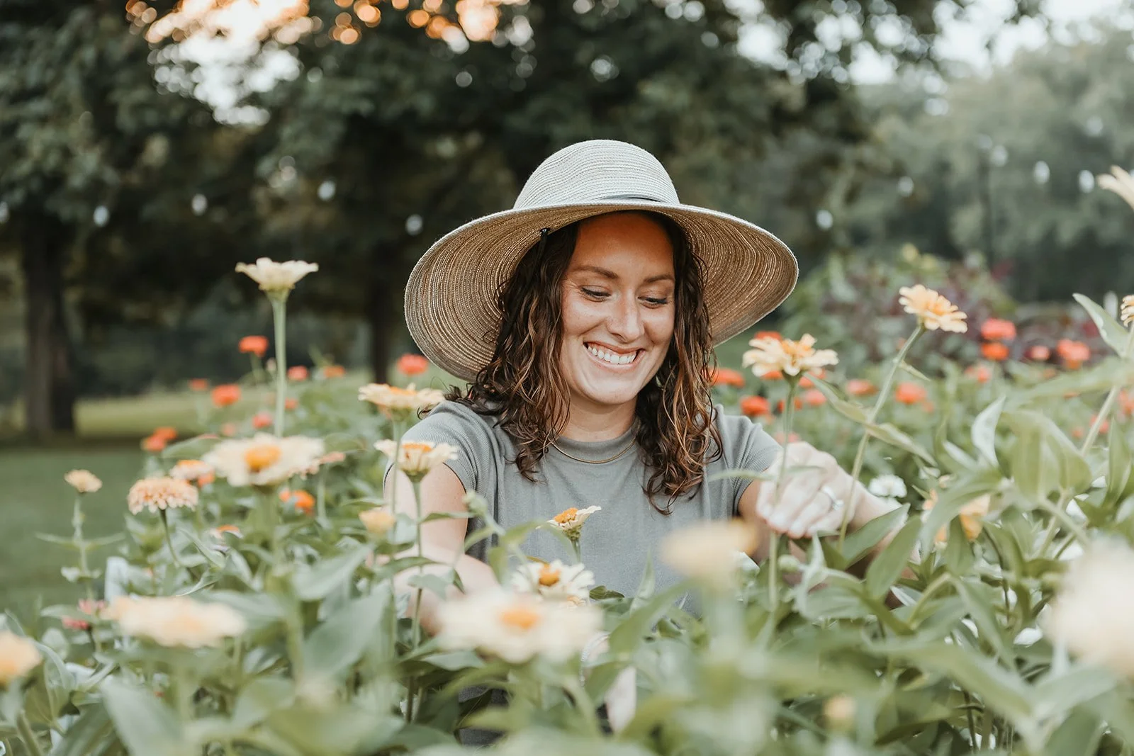 A woman smiling and wearing a large sun hat, standing among colorful flowers in a garden during the daytime.