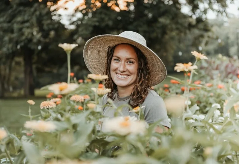 A woman smiling in a sunlit flower garden, wearing a wide-brimmed straw hat and a light gray shirt.