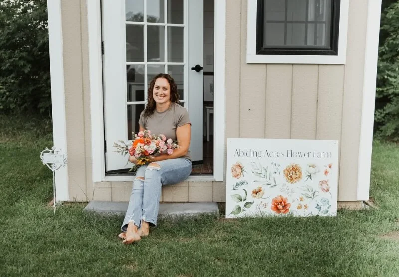 A woman with brown hair wearing a gray t-shirt and ripped jeans sitting on a small concrete step outside a small building, holding a colorful bouquet of flowers, with a floral-themed sign reading 'Abiding Acres Flower Farm' beside her on the grass.