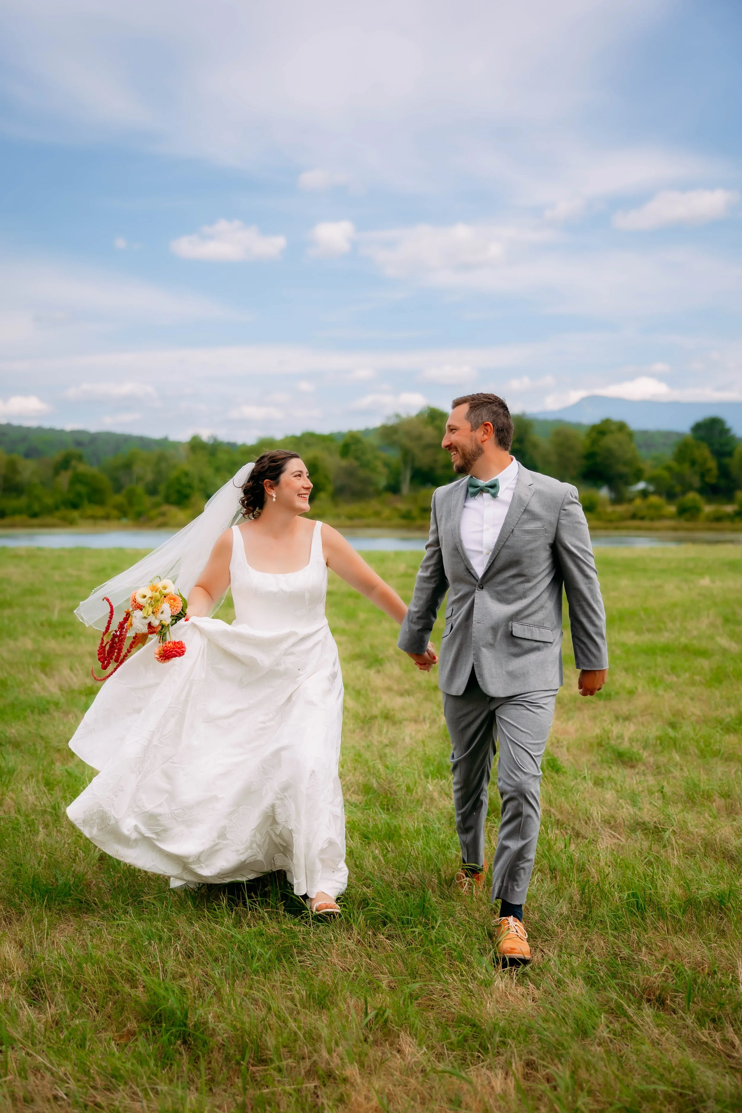 A bride and groom walking hand in hand through a grassy field outdoors, smiling at each other on their wedding day.