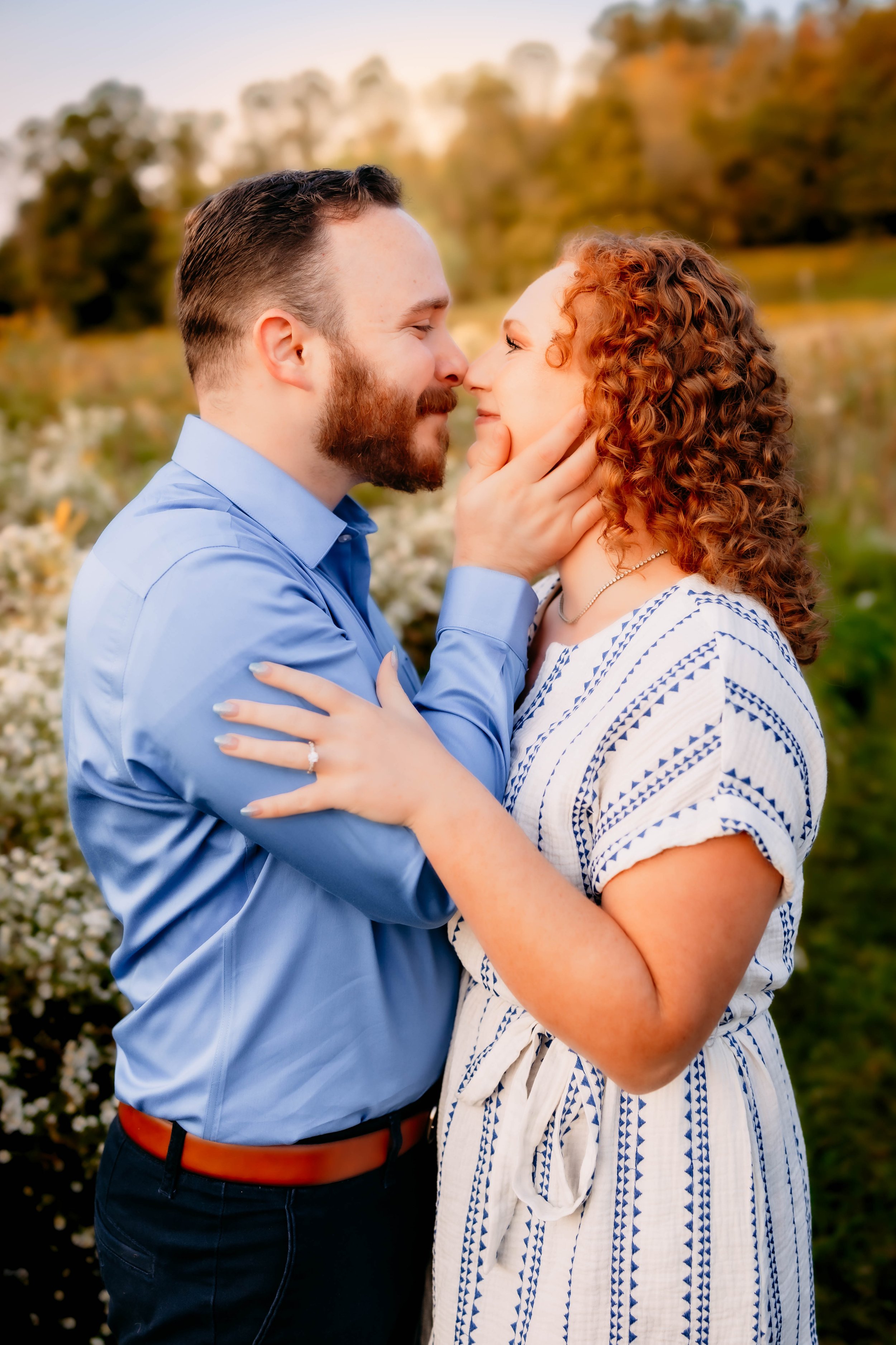 A couple sharing a romantic moment outdoors during golden hour, close to a lake or pond, with trees and flowers in the background.