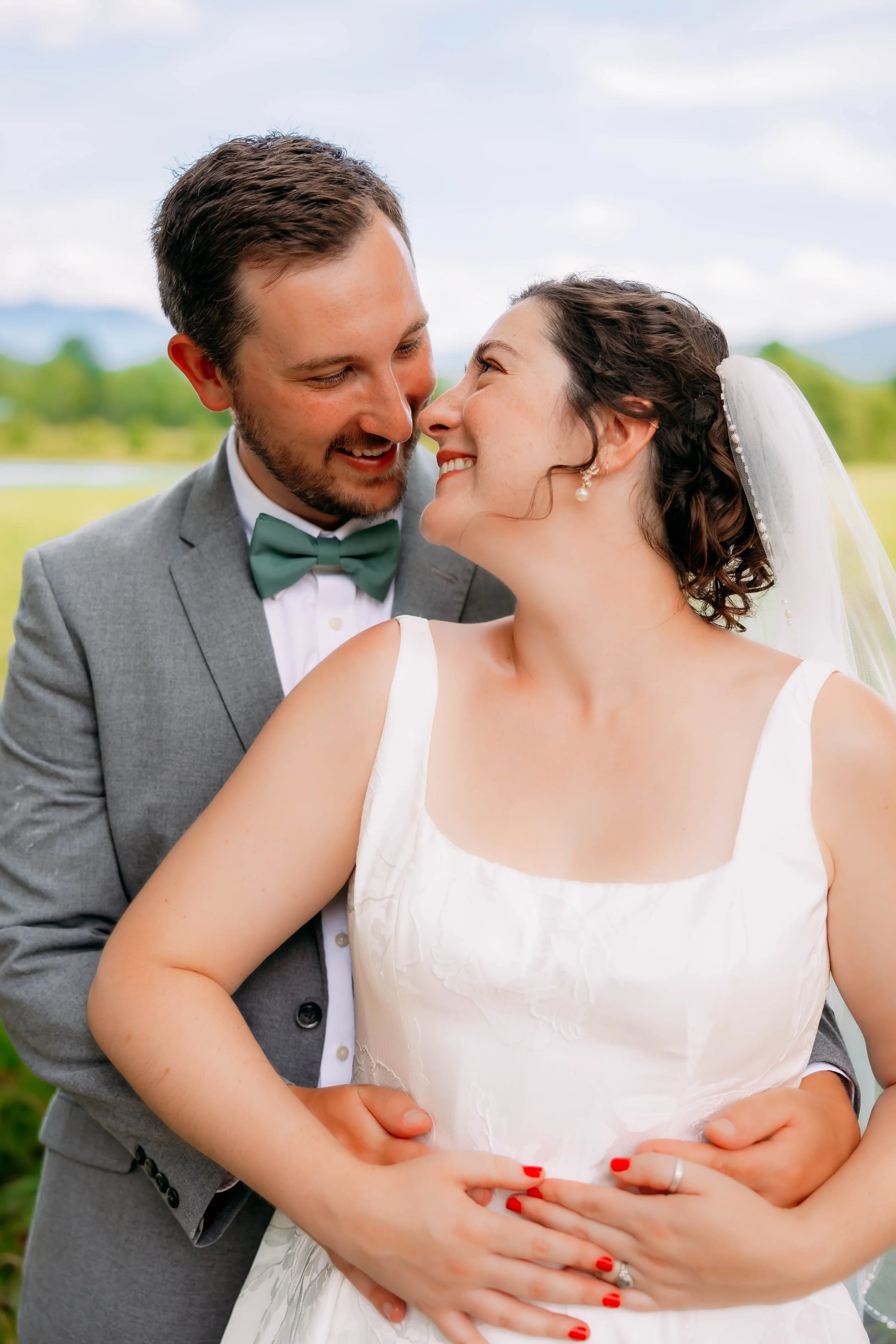 A bride and groom are on their wedding day outdoors, smiling and looking at each other. The groom is in a gray suit with a green bow tie, and the bride is in a white wedding dress with a veil and pearl earrings. They are holding hands in front of her