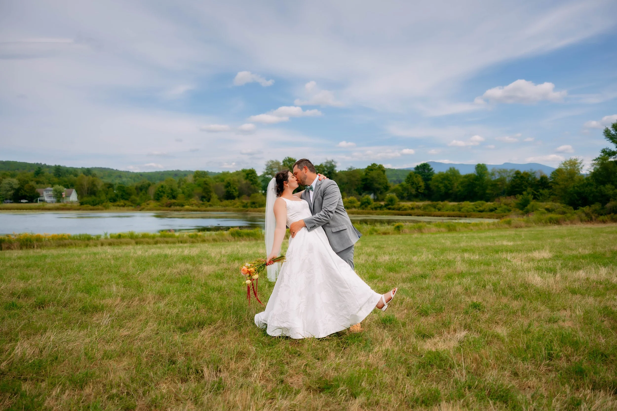 A bride and groom sharing a kiss outdoors on a grassy field, with a lake and green trees in the background under a partly cloudy sky.