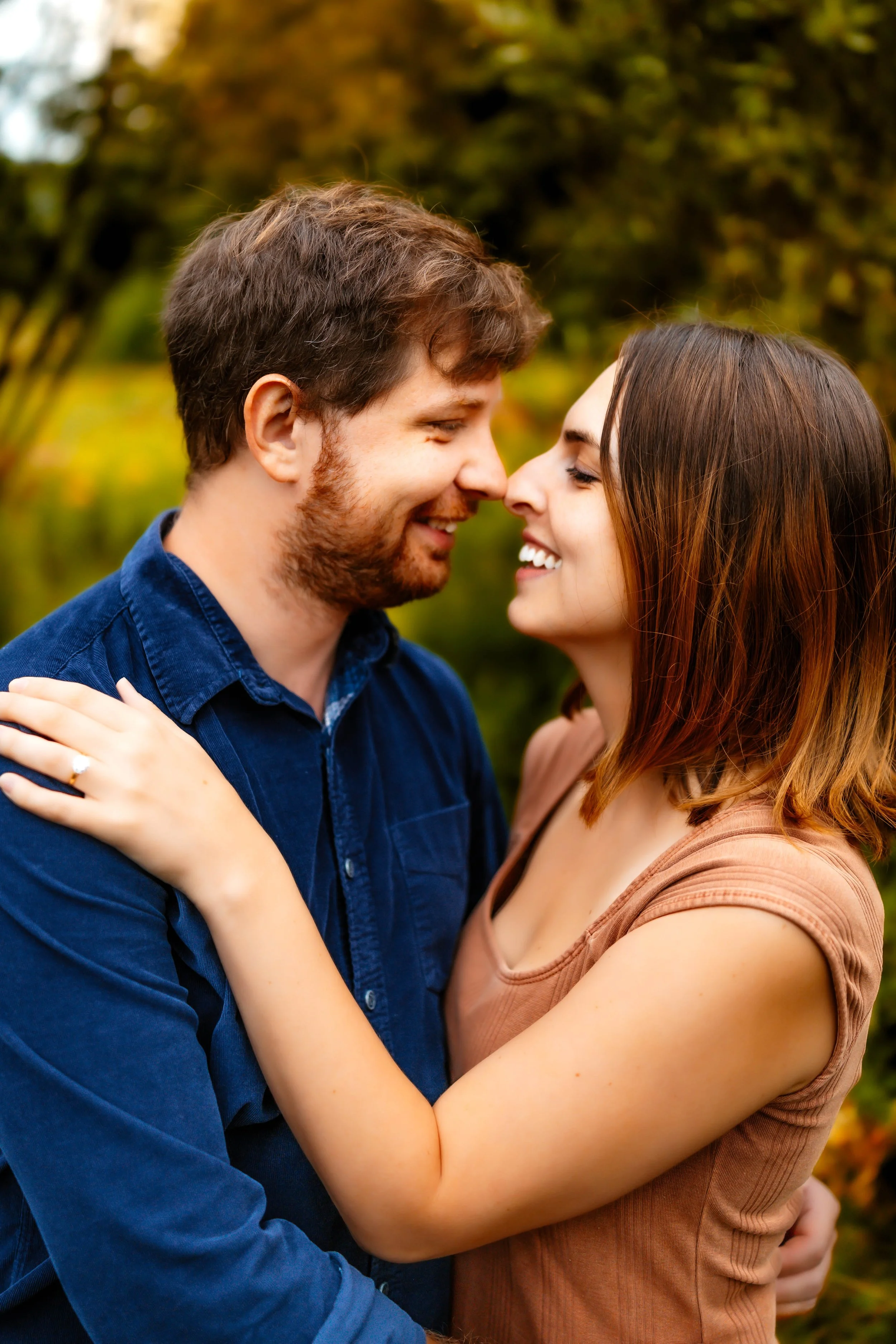 A couple standing closely, smiling, and looking into each other's eyes outdoors with trees and greenery in the background.