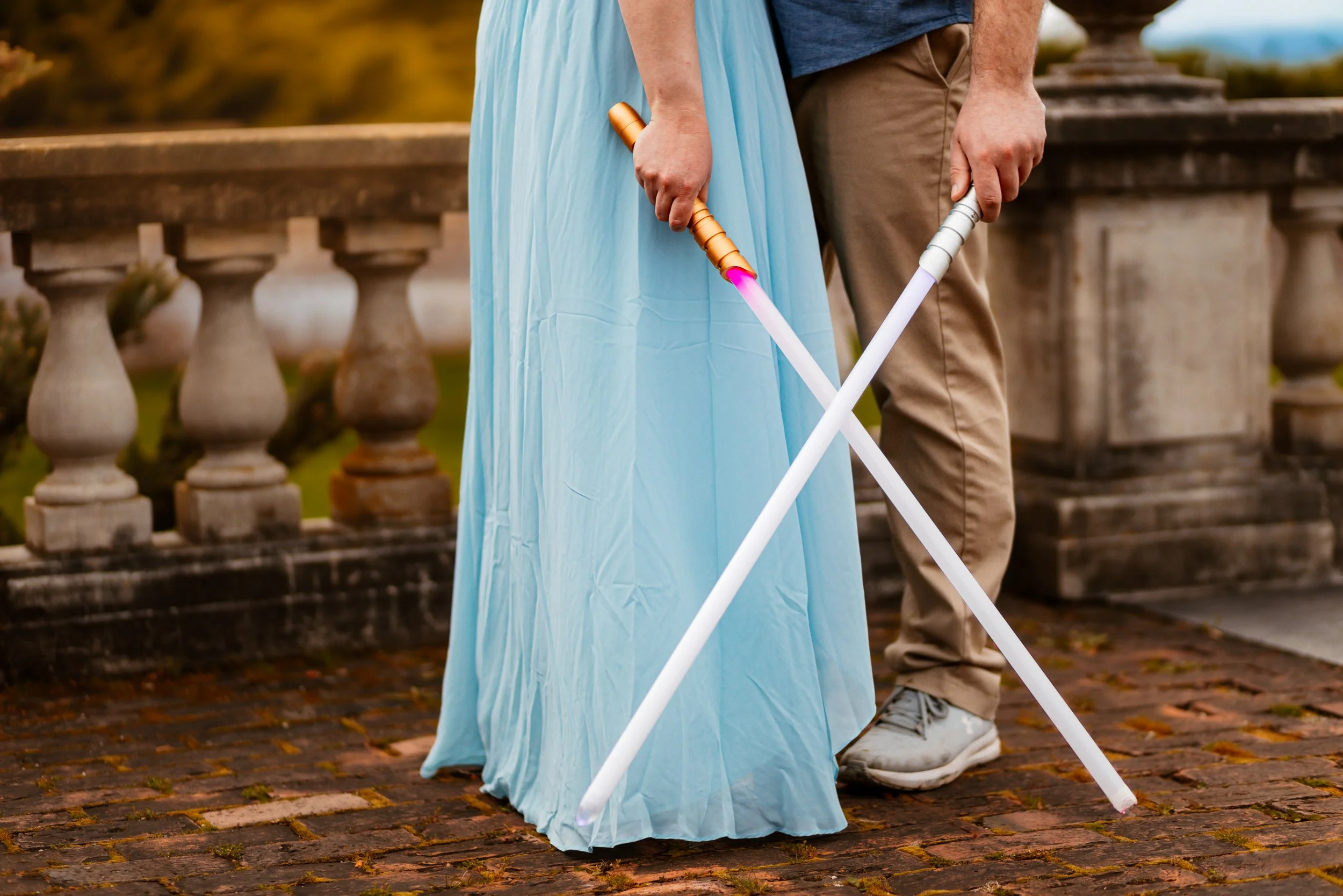 Two people dressed as characters from Star Wars, holding lightsabers and standing outdoors on a brick pathway with a stone railing in the background.