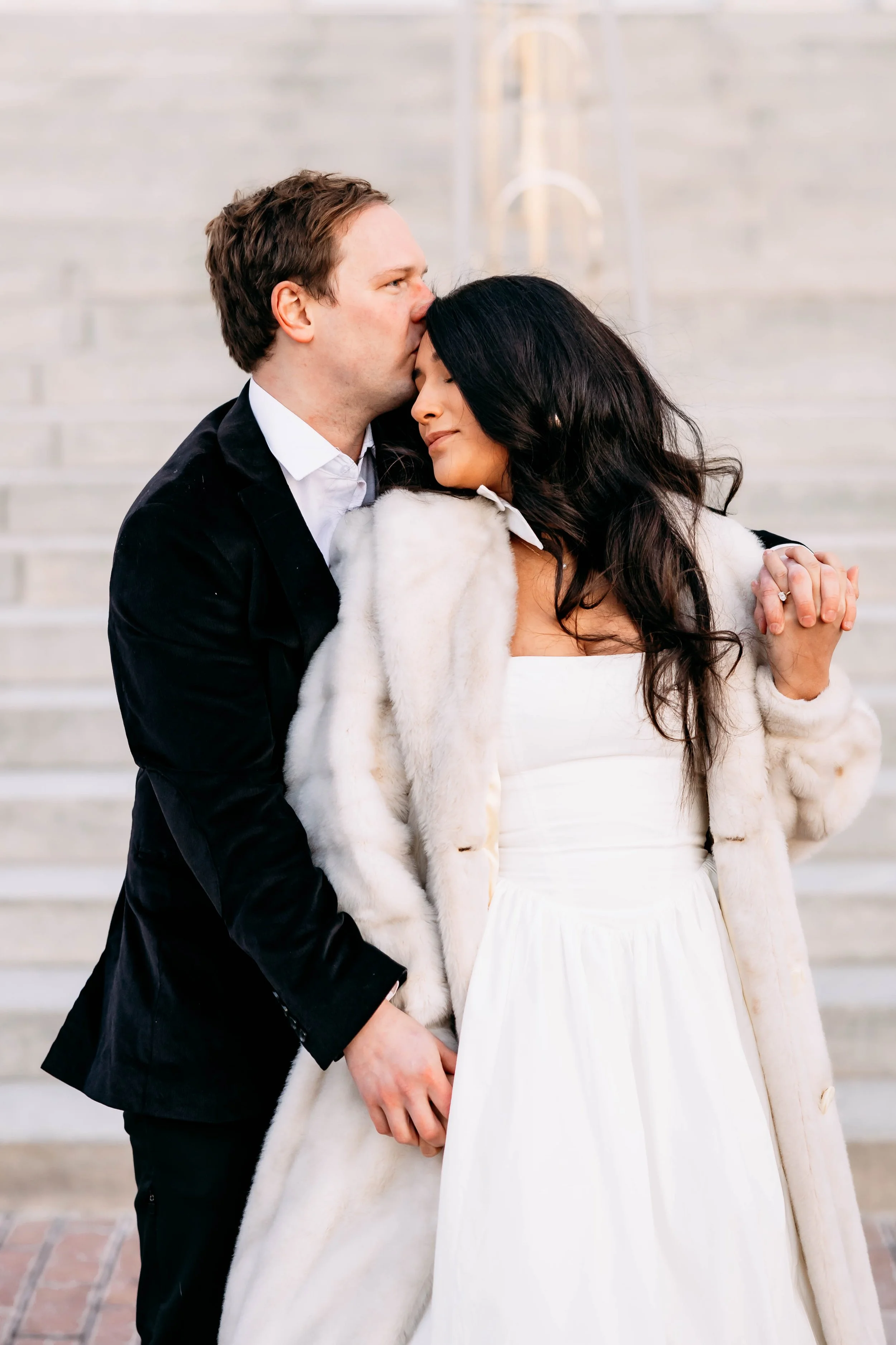 A man tenderly kisses a woman's forehead as she smiles with closed eyes, both dressed in formal attire, holding hands, standing on stairs.