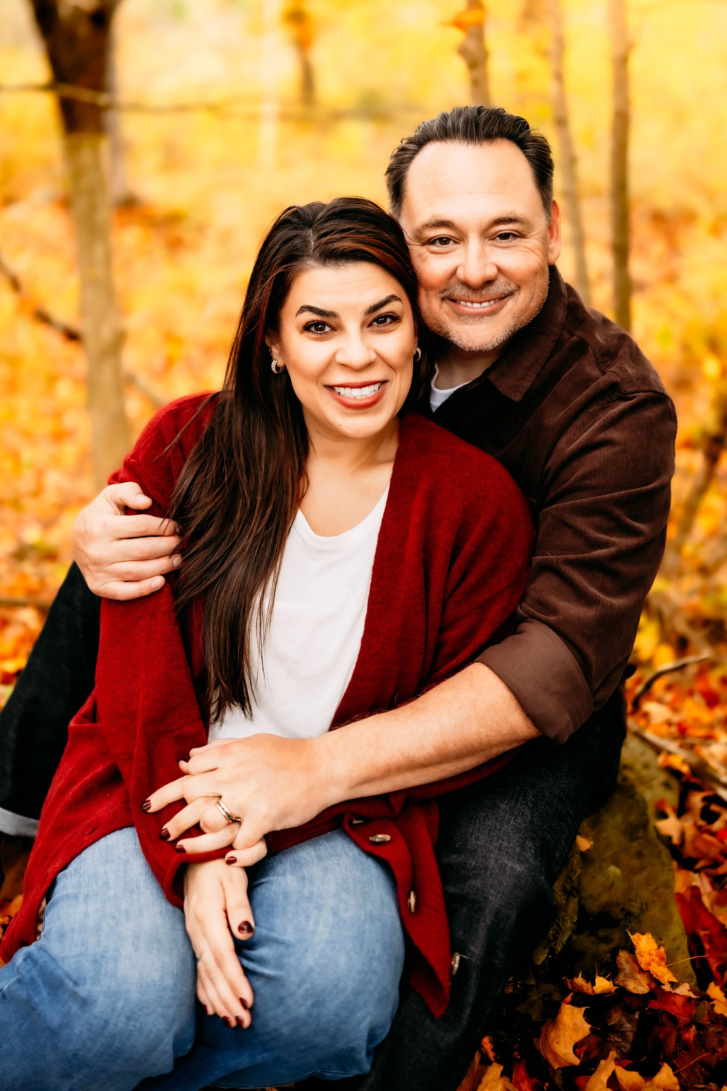 A smiling couple sitting outdoors in an autumnal forest, surrounded by fallen leaves, embracing each other.