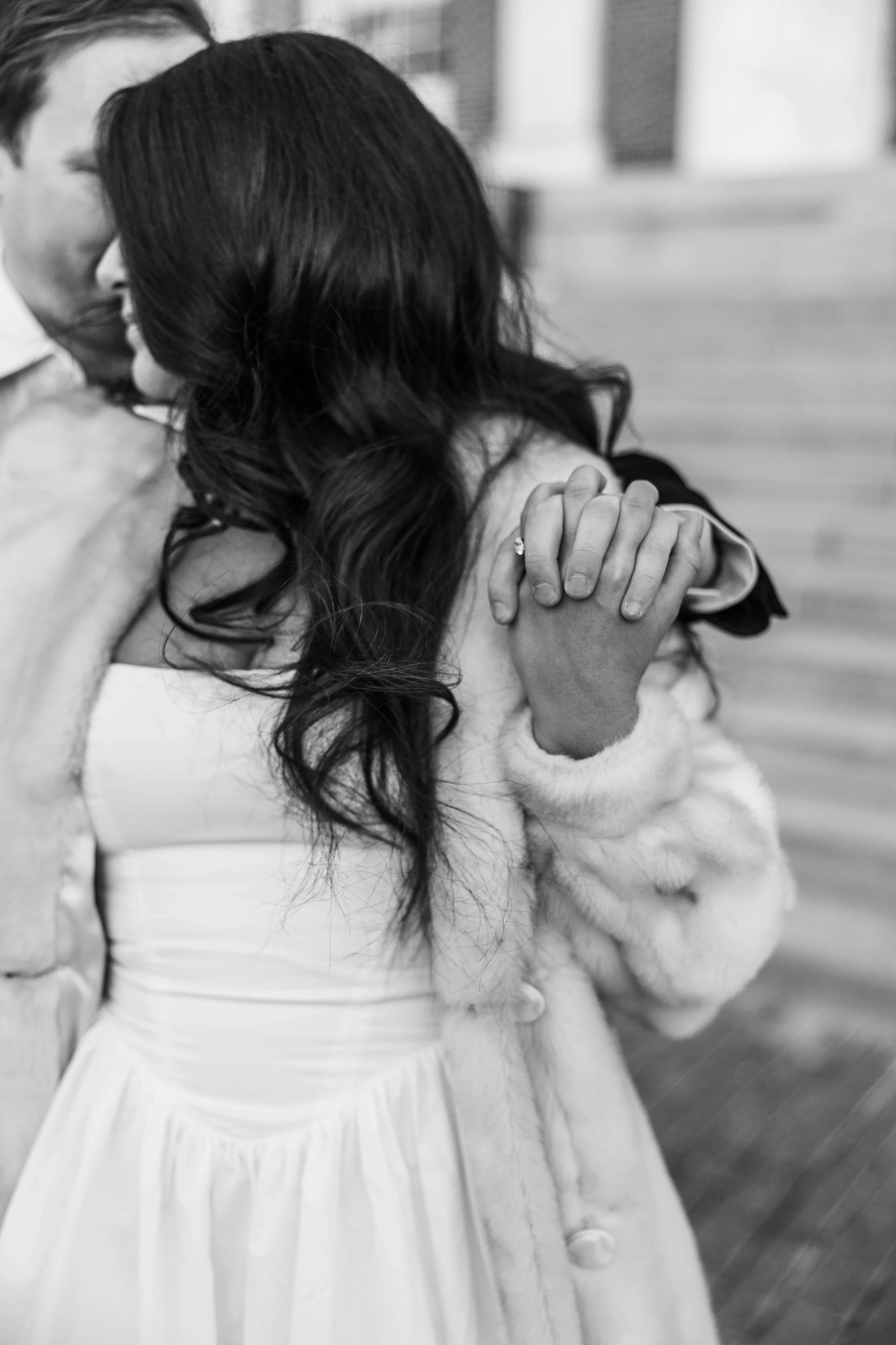 A couple embracing closely, holding hands, with the woman wearing a white dress and fur coat, in black and white.