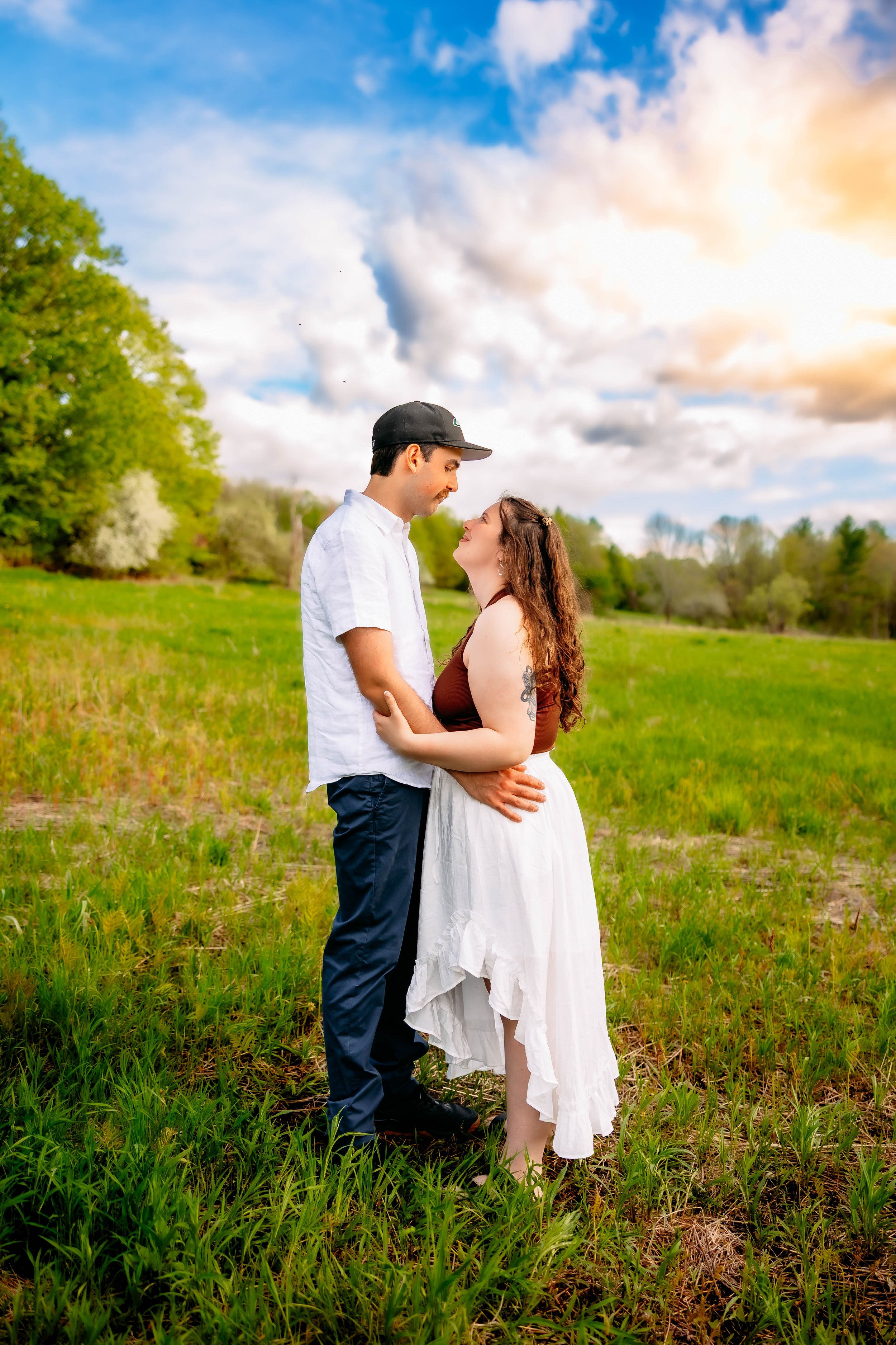 A couple standing close together in a green field, gazing at each other, with trees and a partly cloudy sky in the background during sunset.