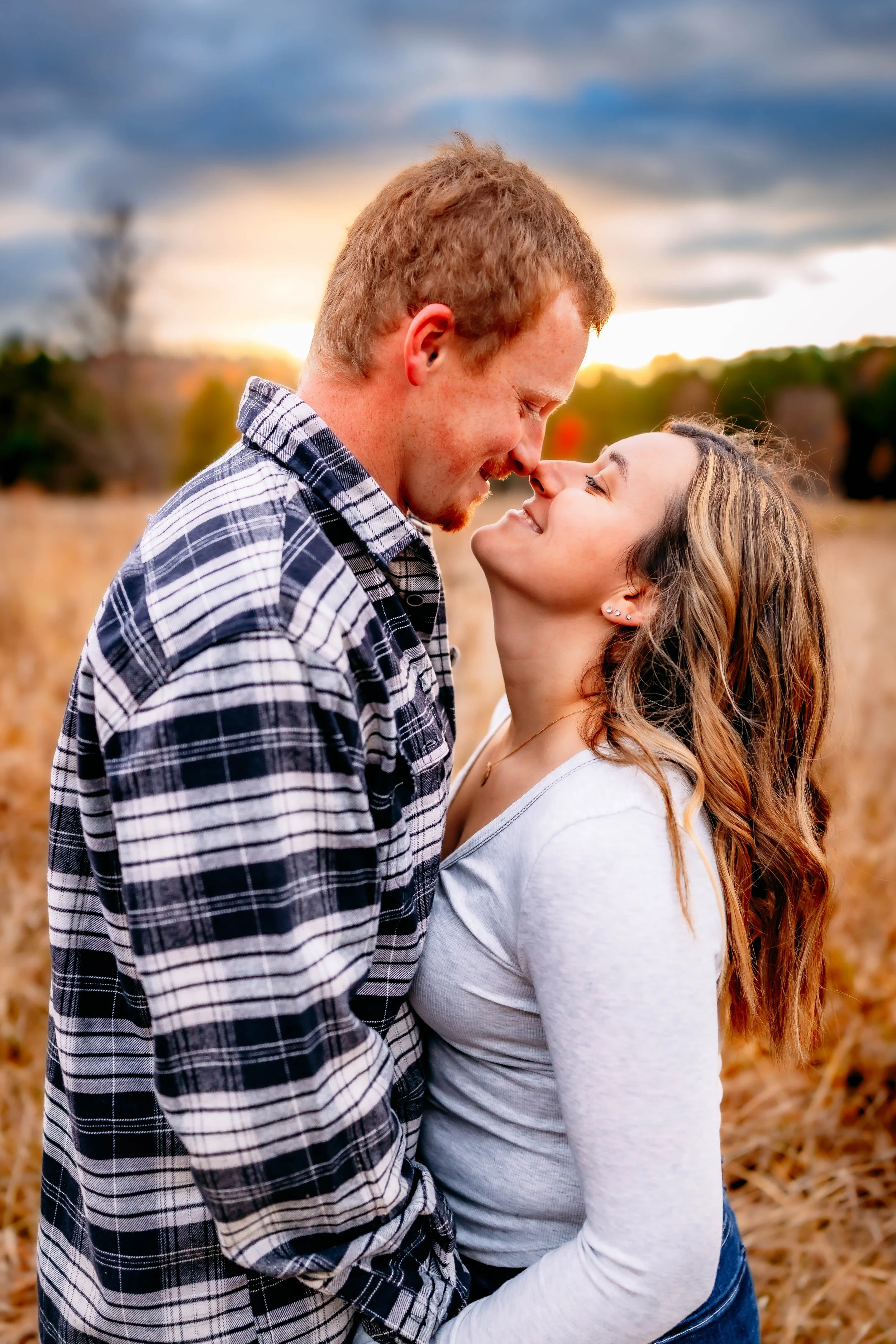A couple standing close together outdoors during sunset, smiling and leaning their foreheads together.