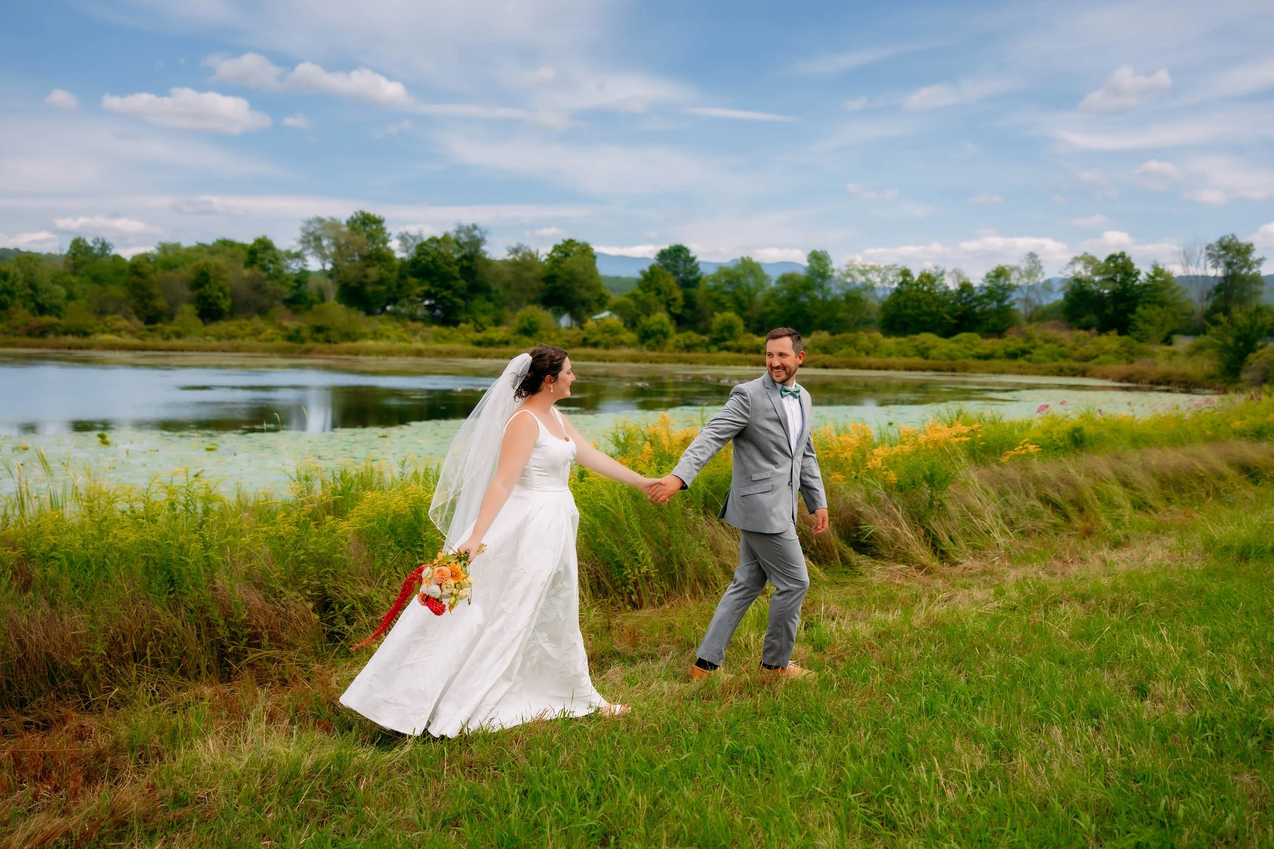 Bride and groom holding hands in a grassy field by a lake with trees and a cloudy sky in the background.