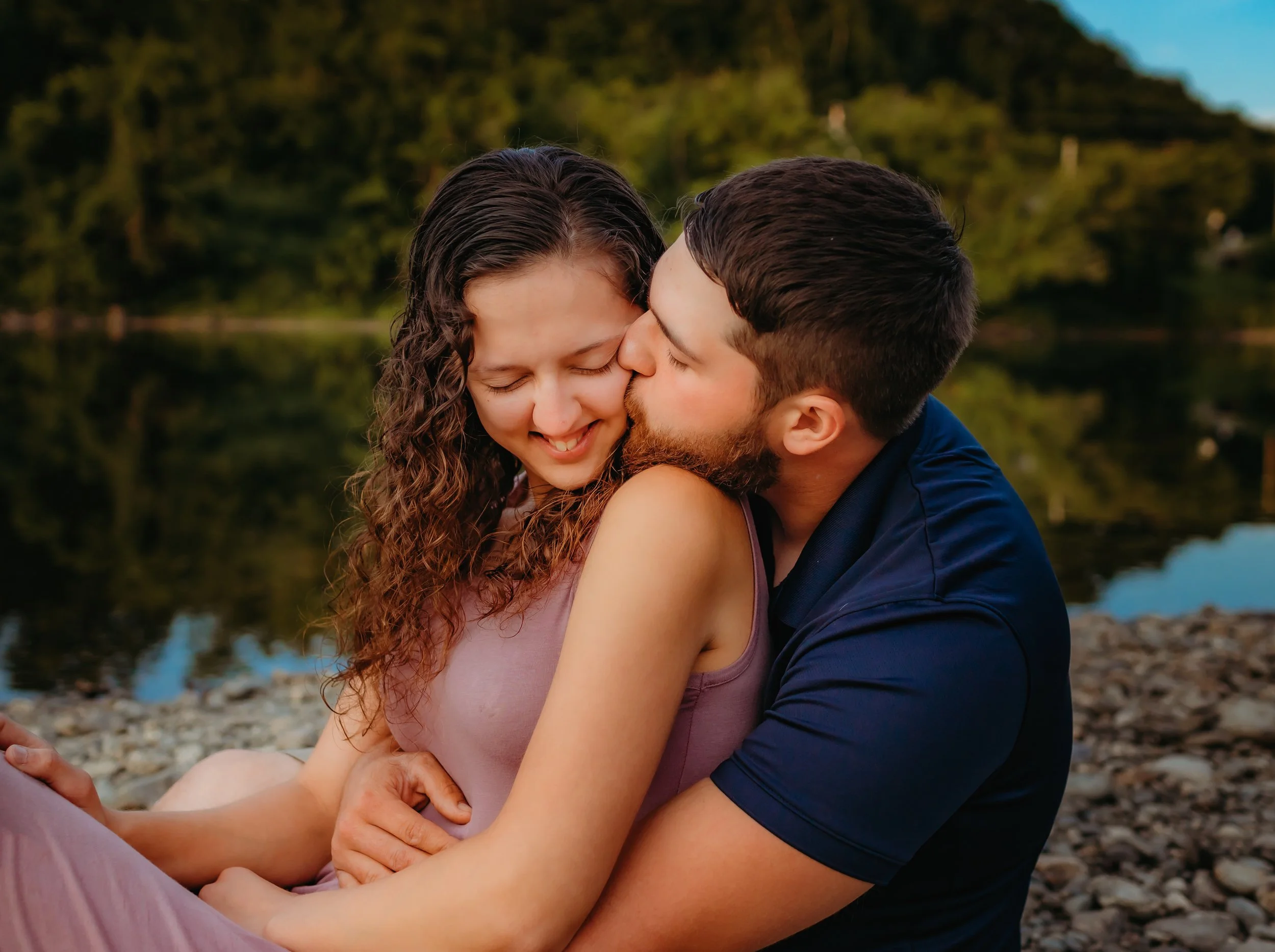 A man kissing a woman on the cheek near a body of water with a forested landscape in the background.