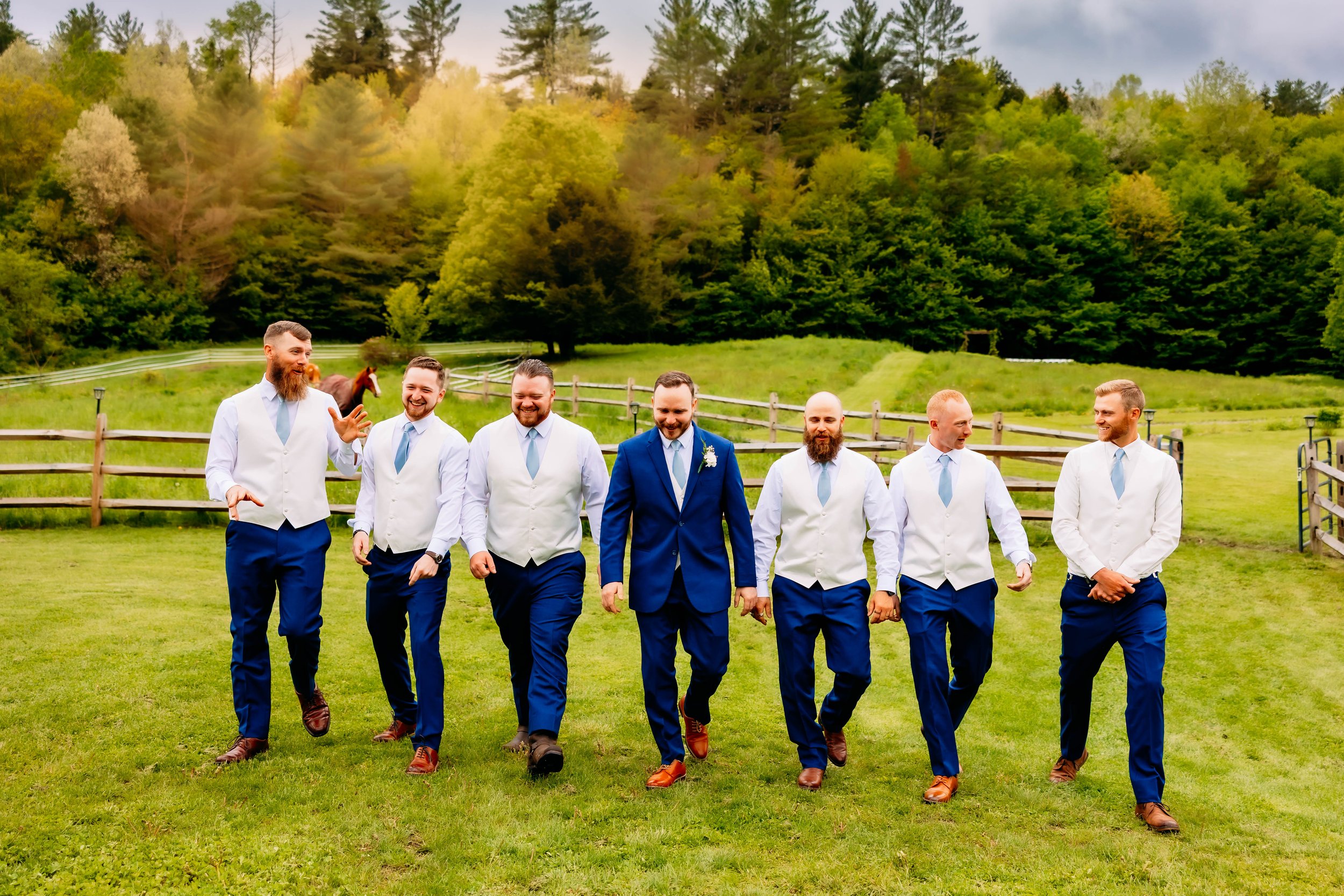 A group of seven men dressed in formal wedding attire walking outdoors on a grassy field, with trees and a wooden fence in the background.