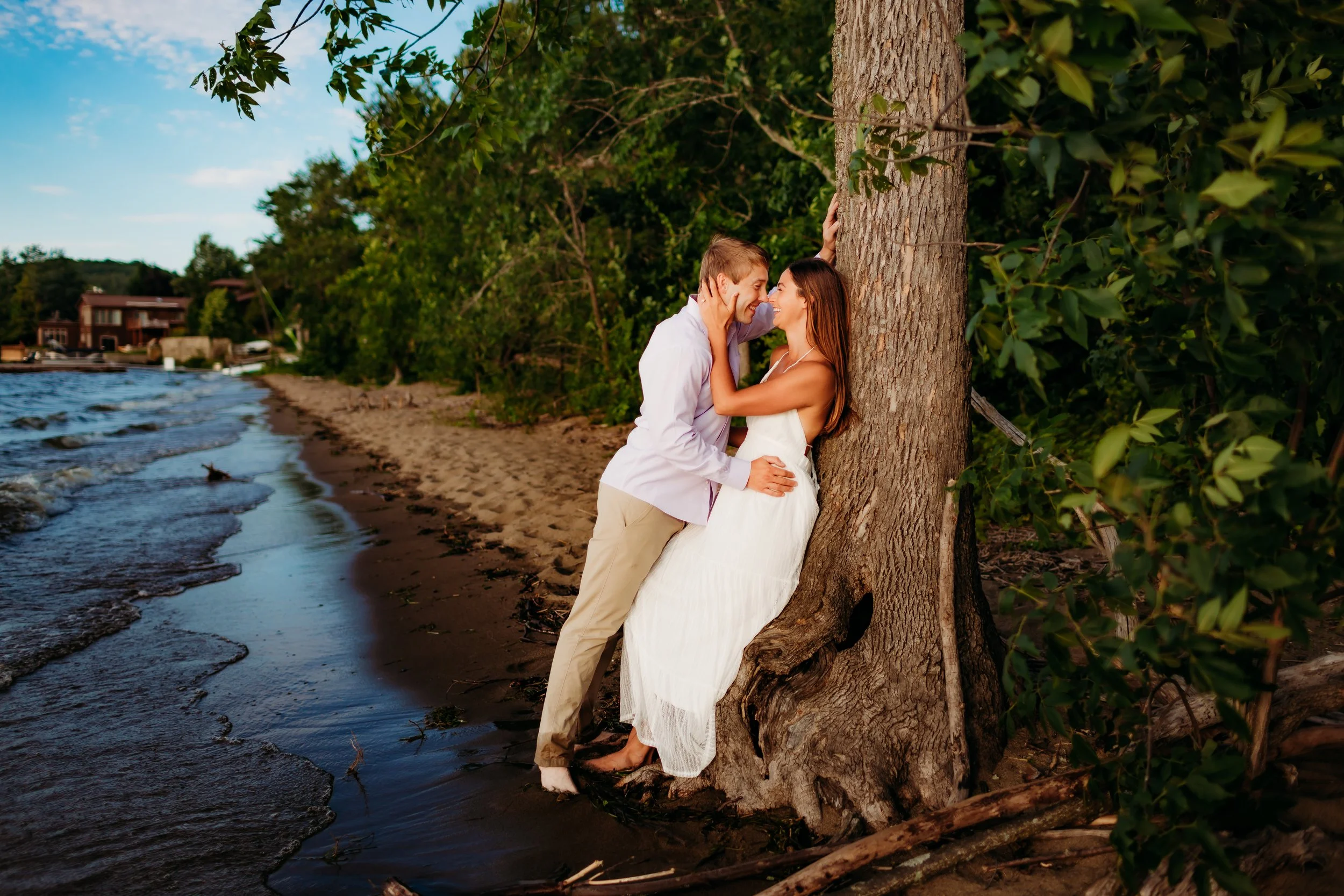 A couple stands on a beach, leaning against a large tree, facing each other with their noses touching, enjoying an intimate moment by the water, surrounded by greenery and houses in the background.
