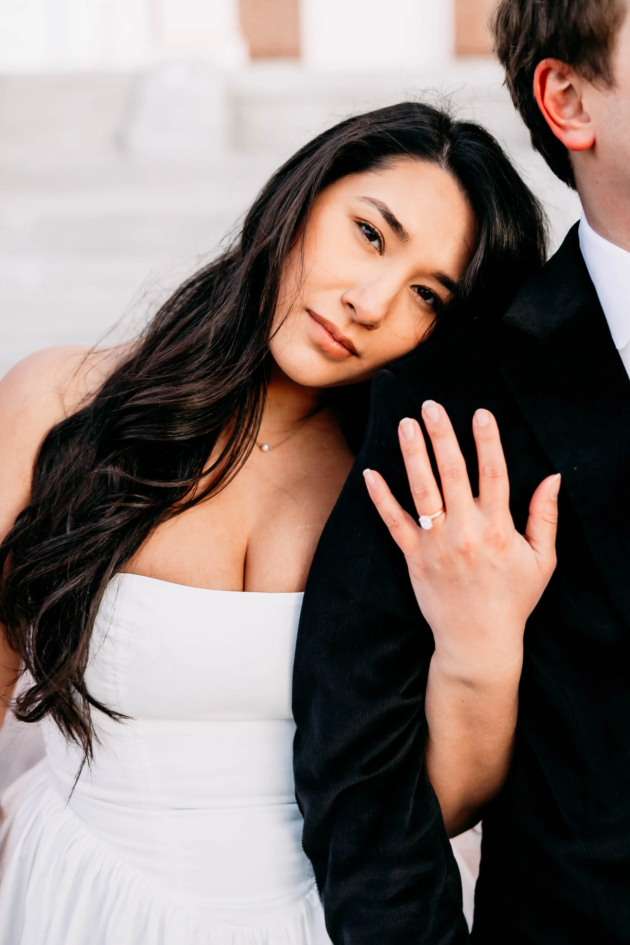 A woman with long dark hair resting her head on a man's shoulder. She is wearing a white dress and a ring on her left hand, with her hand gently placed on his shoulder. The man is wearing a black suit with a white shirt. The background is blurred.