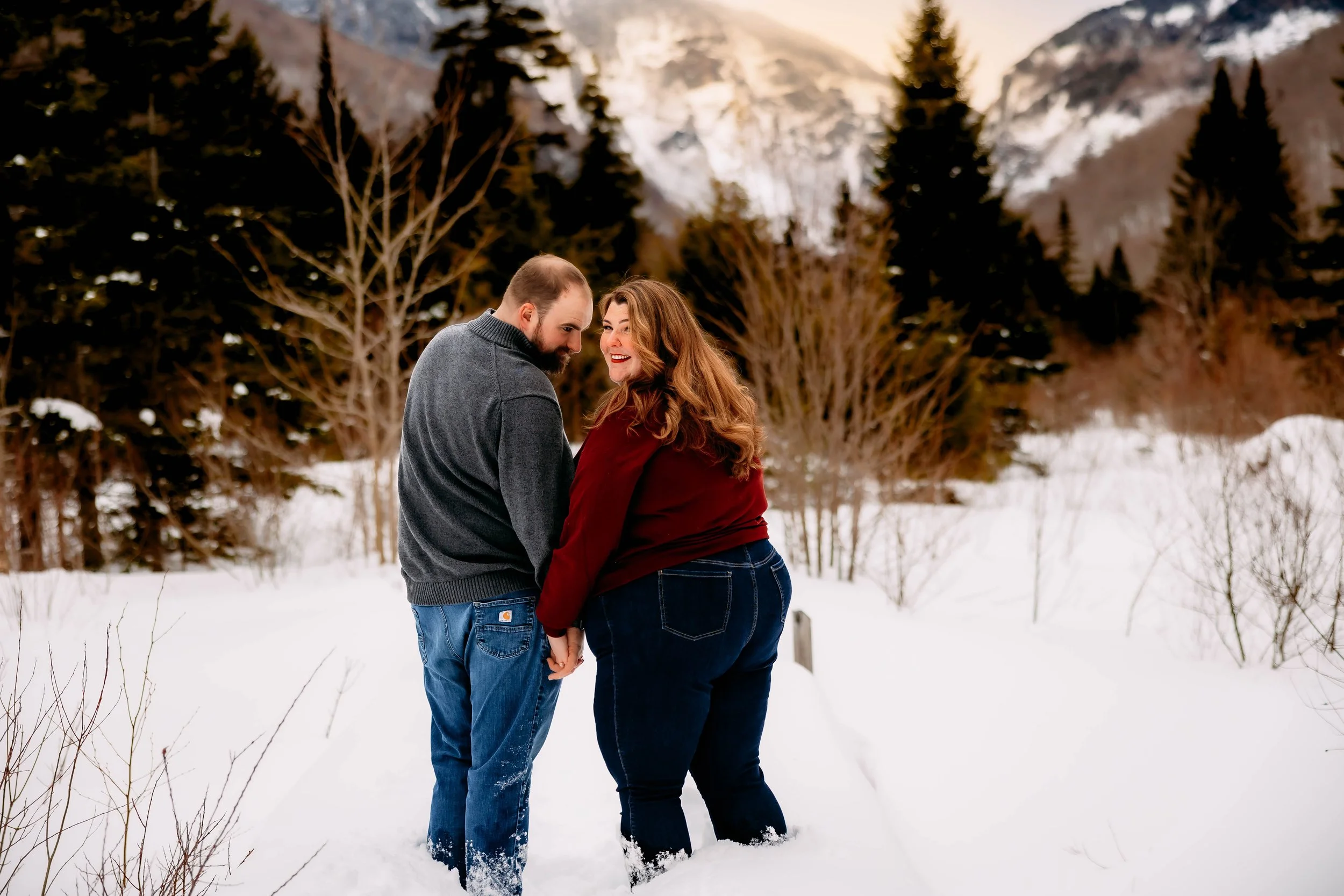 A couple holding hands and smiling at each other in a snowy landscape with pine trees and mountain background.