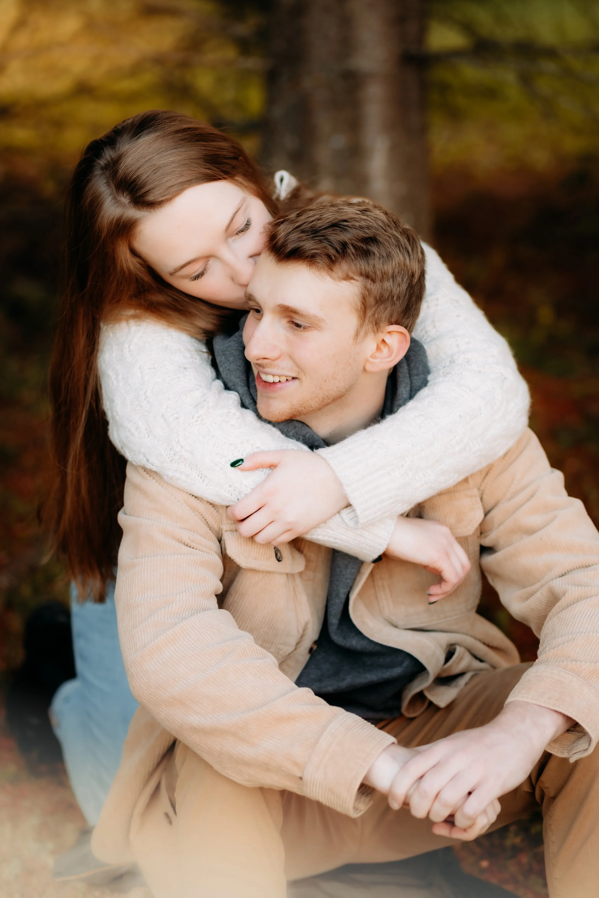 A young couple sitting outdoors in a wooded area, the woman hugging the man from behind and kissing his head.