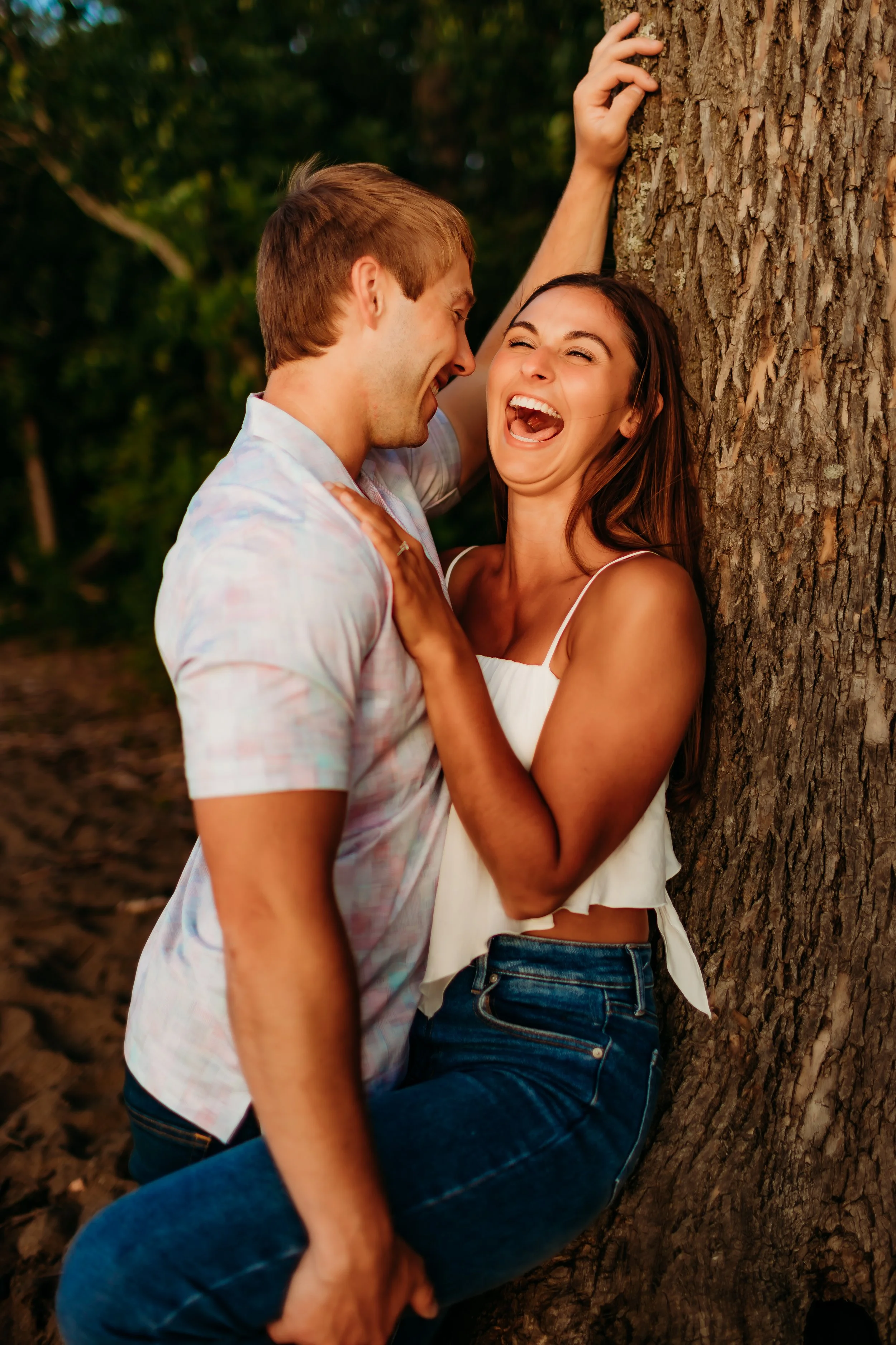 A couple laughing and touching each other while leaning against a tree in a forest setting.
