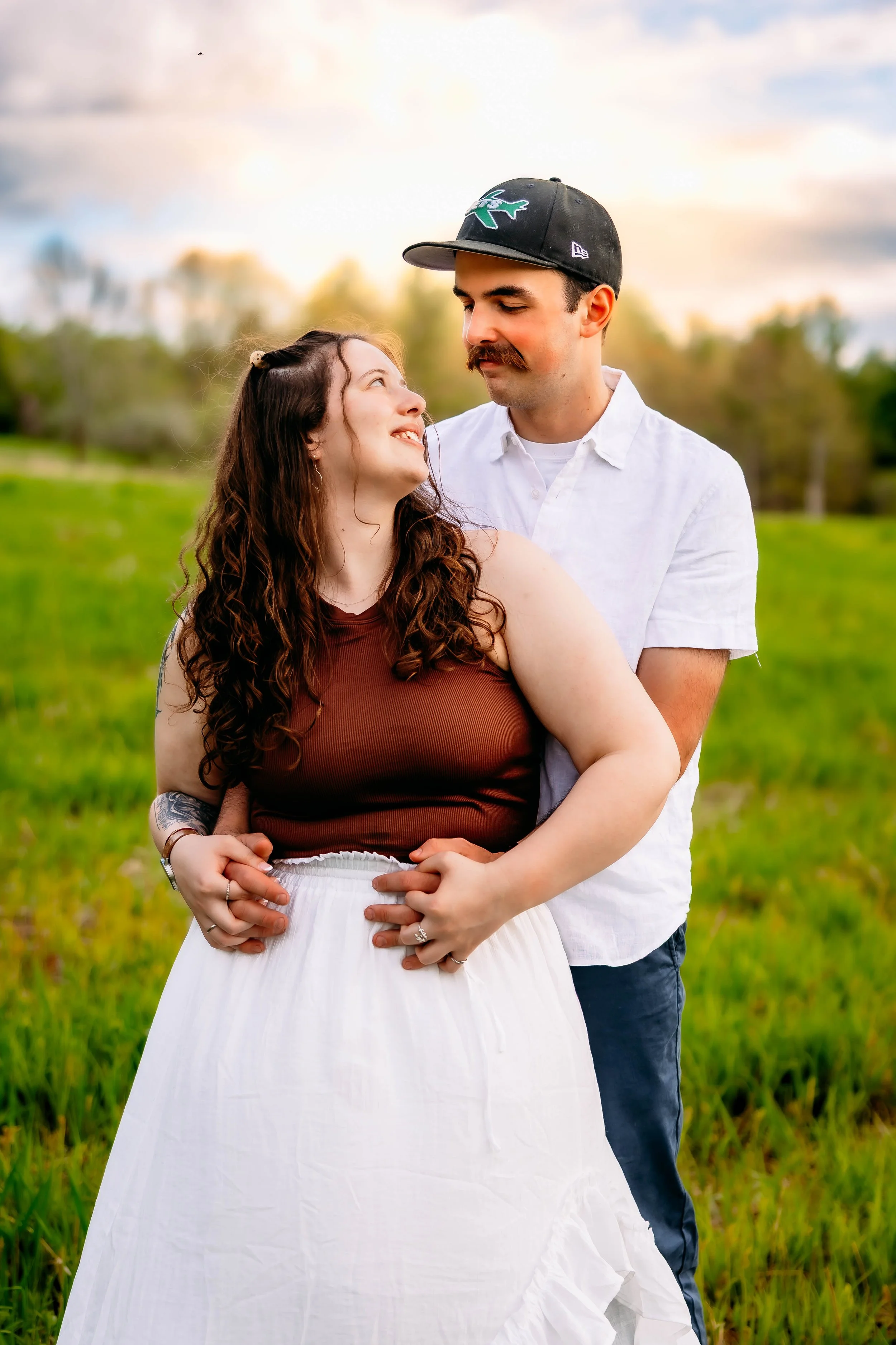 A couple embracing outdoors in a grassy field at sunset, looking into each other's eyes.