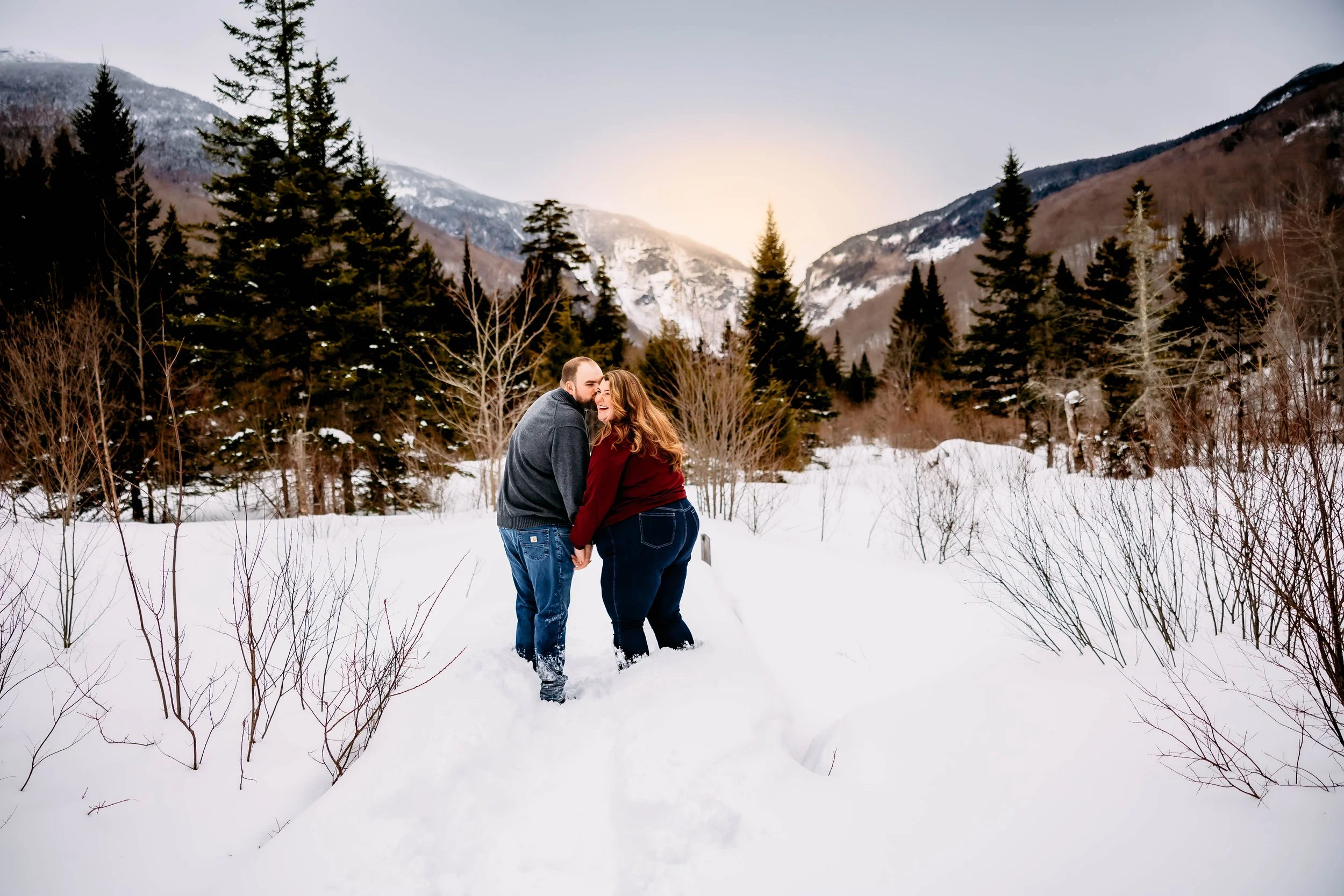 A couple holding hands and smiling in a snowy landscape with evergreen trees and mountains in the background.