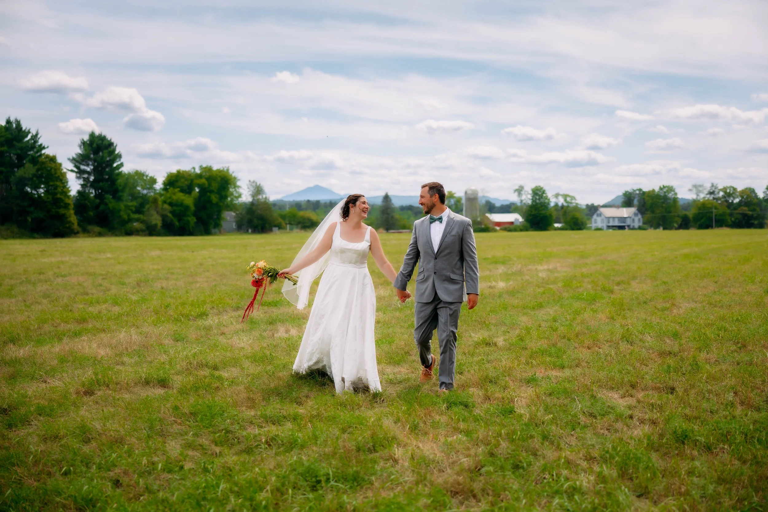 A newlywed couple holding hands and walking through a grassy field on a cloudy day, with trees, houses, and distant mountains in the background.