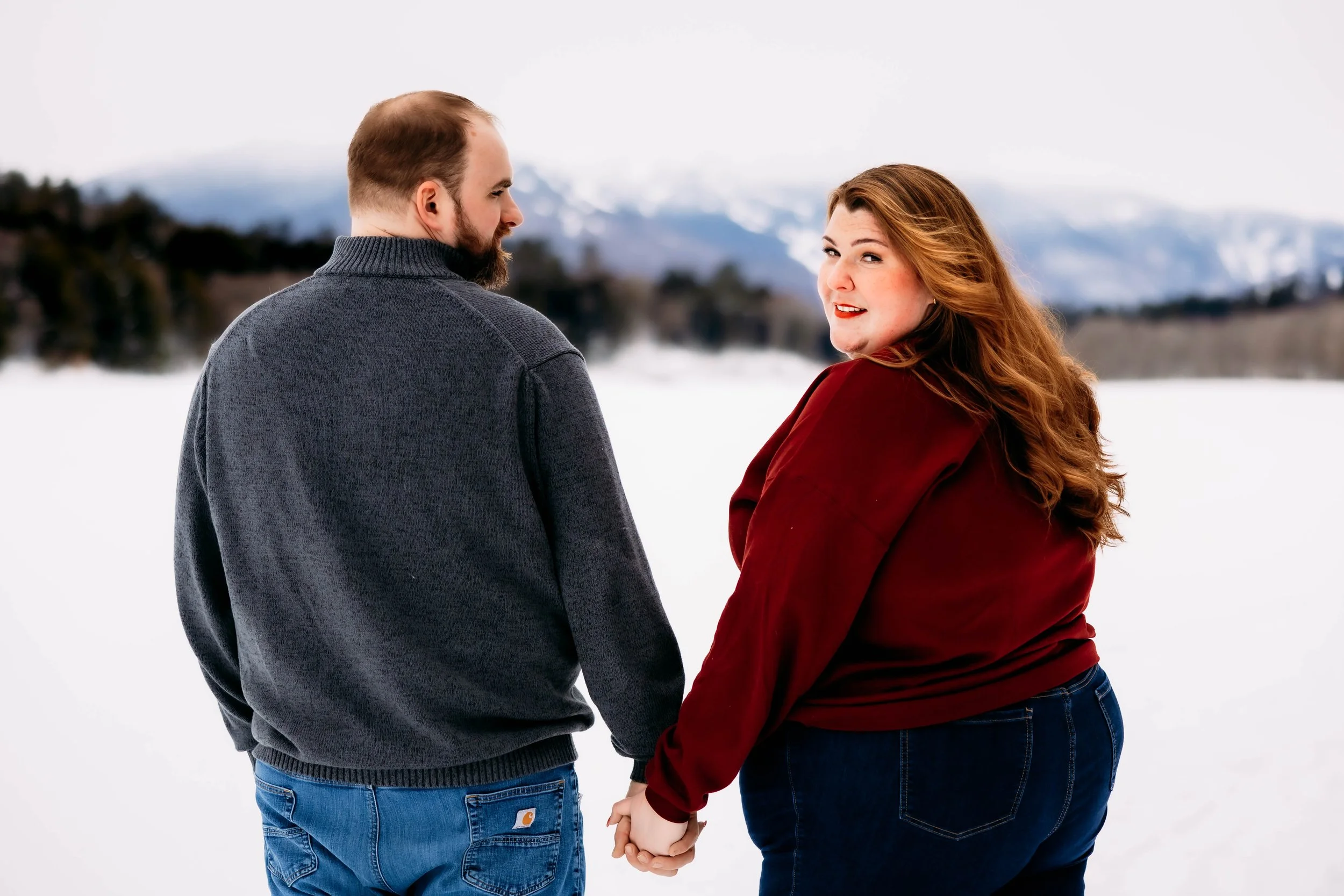 A couple holding hands walking in a snowy landscape with mountains in the background, one man with a beard wearing a gray sweater and jeans, and a woman with long red hair wearing a red jacket and jeans.