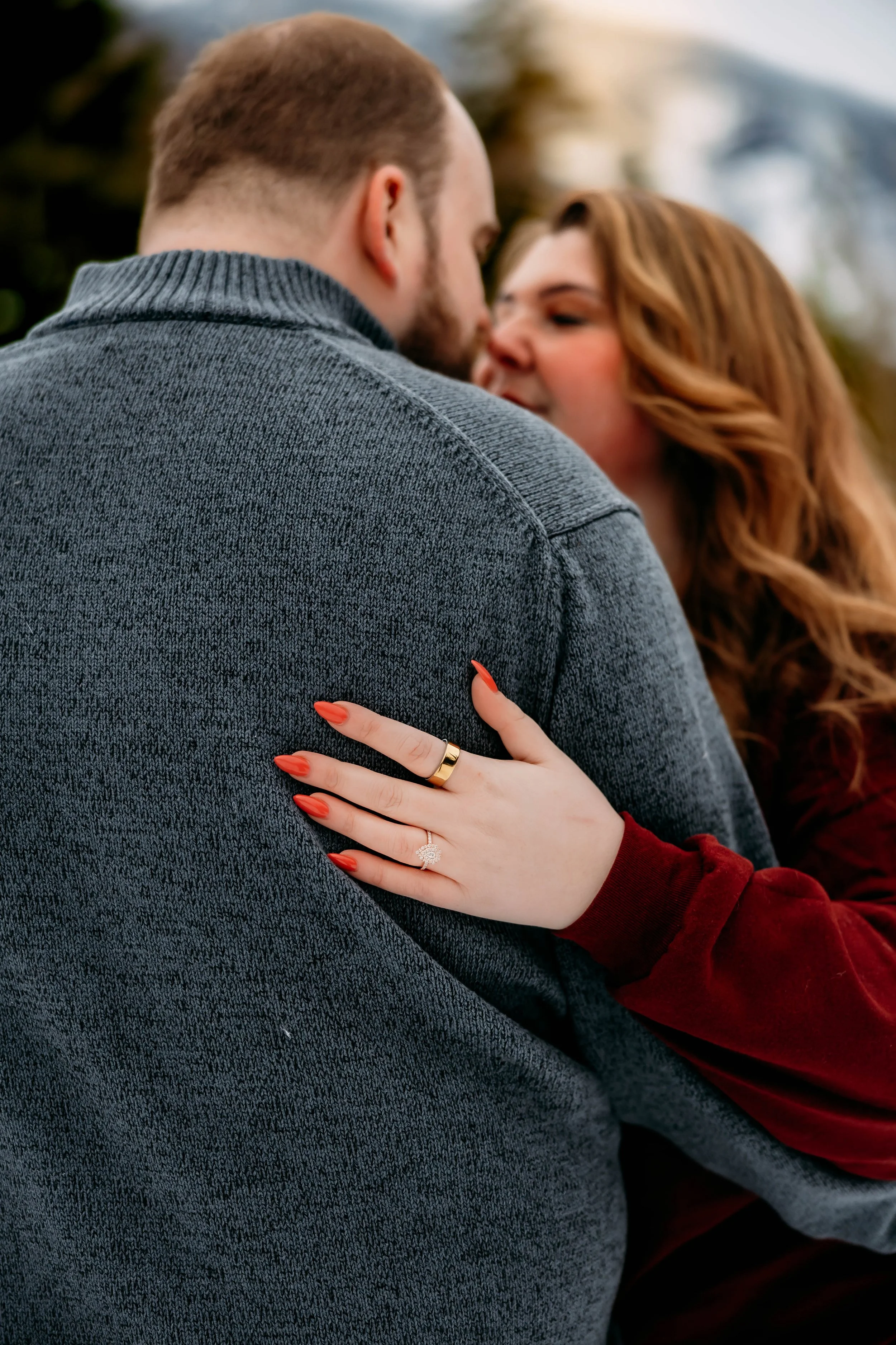 A couple embracing outdoors, the woman's hand with an engagement ring resting on the man's shoulder, her nails painted red, and the woman has wavy red hair.