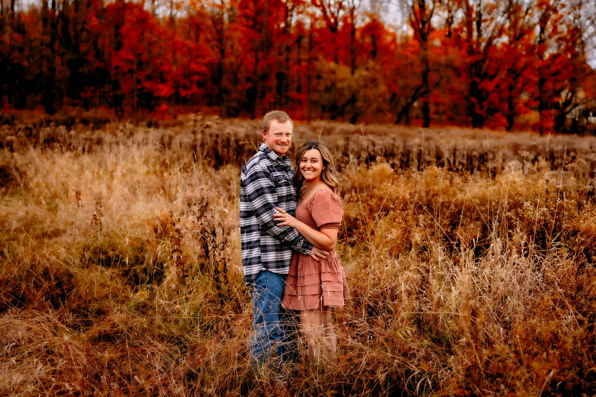 A smiling couple stands in a field of tall, dry grass with colorful autumn trees in the background.