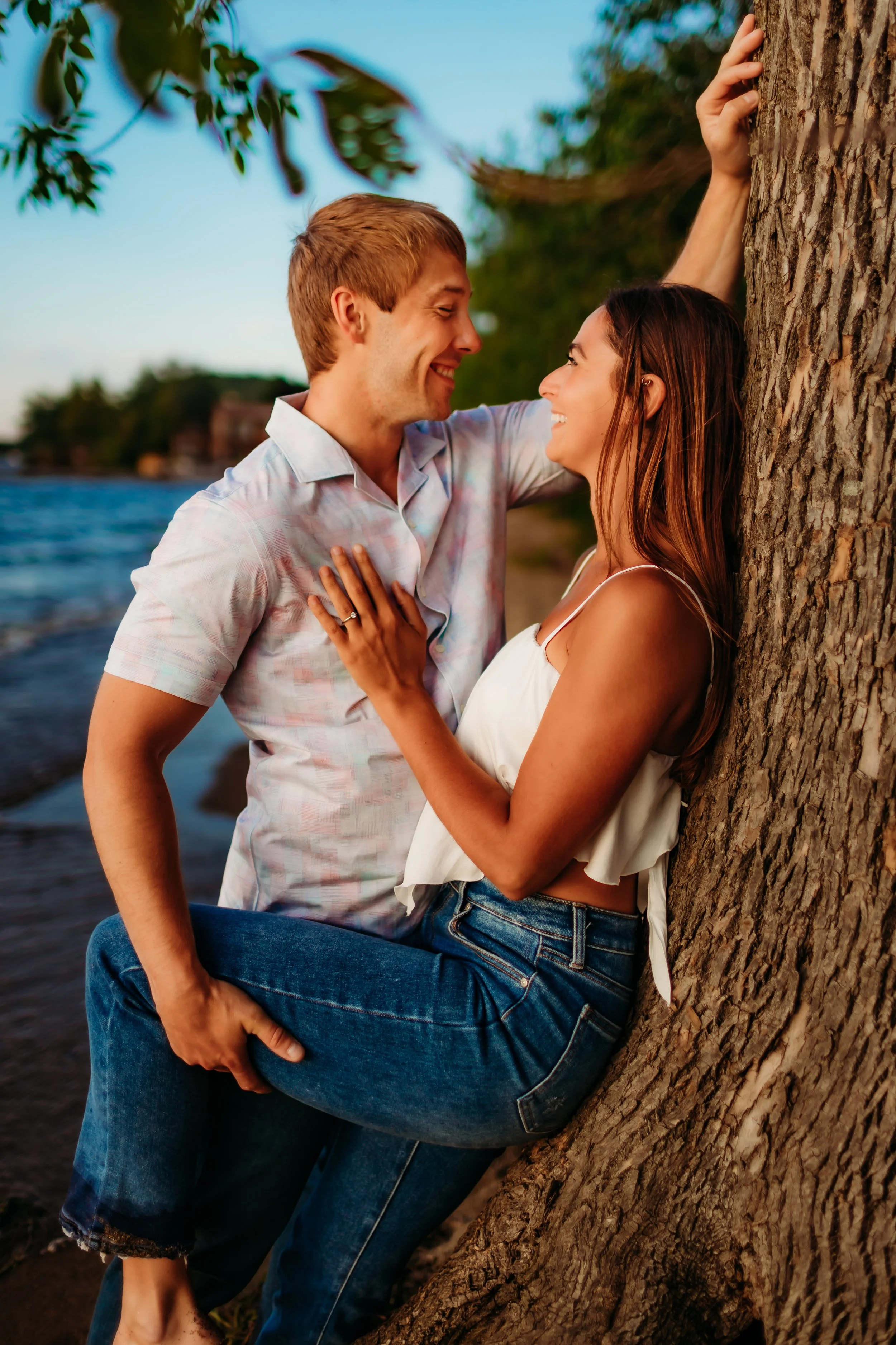 A couple is standing close together near a tree by the water, smiling and looking into each other's eyes during sunset.