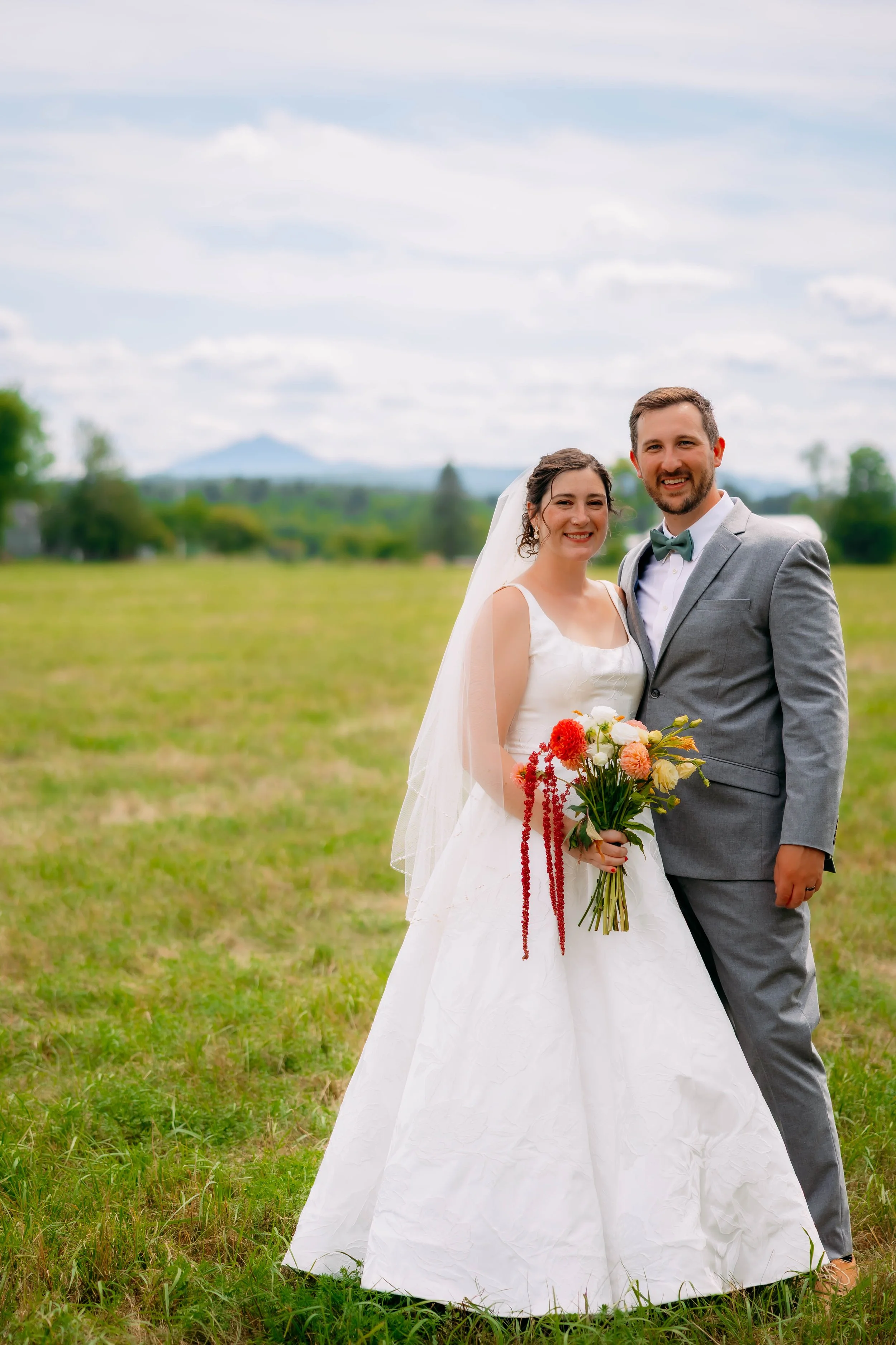 A newlywed couple standing in a grassy field, smiling at the camera. The bride is holding a bouquet of flowers and wearing a white wedding dress with a veil. The groom is wearing a gray suit with a bow tie. The background features a scenic landscape