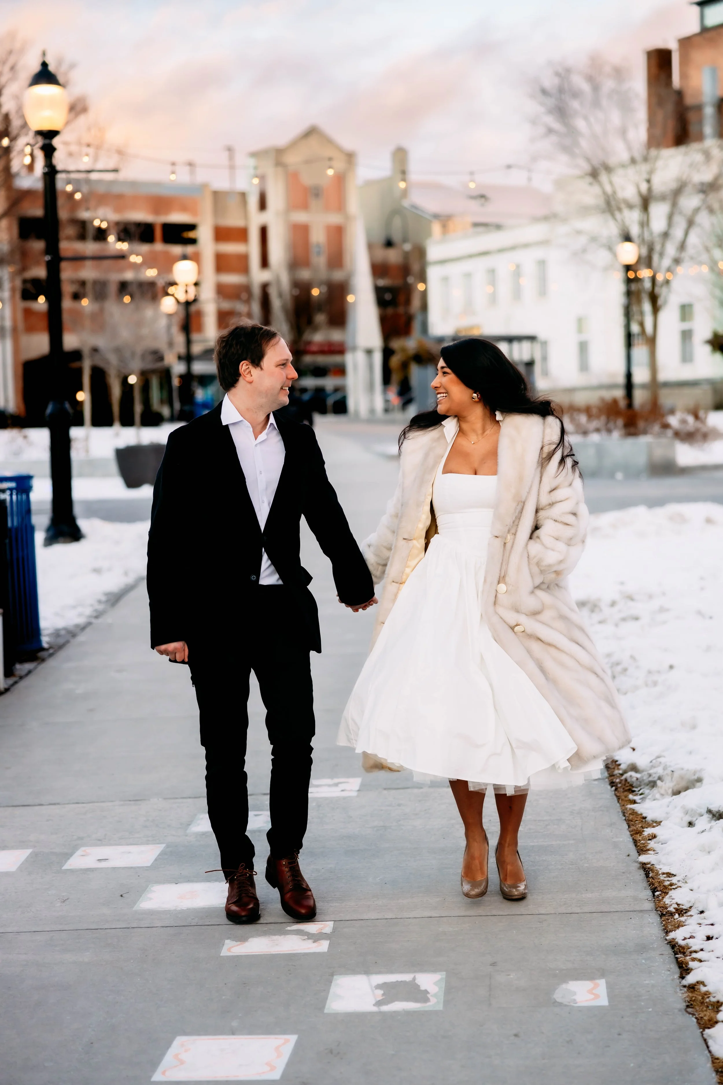 A couple walking hand-in-hand on a snowy sidewalk, dressed for a formal occasion, with city buildings and string lights in the background.
