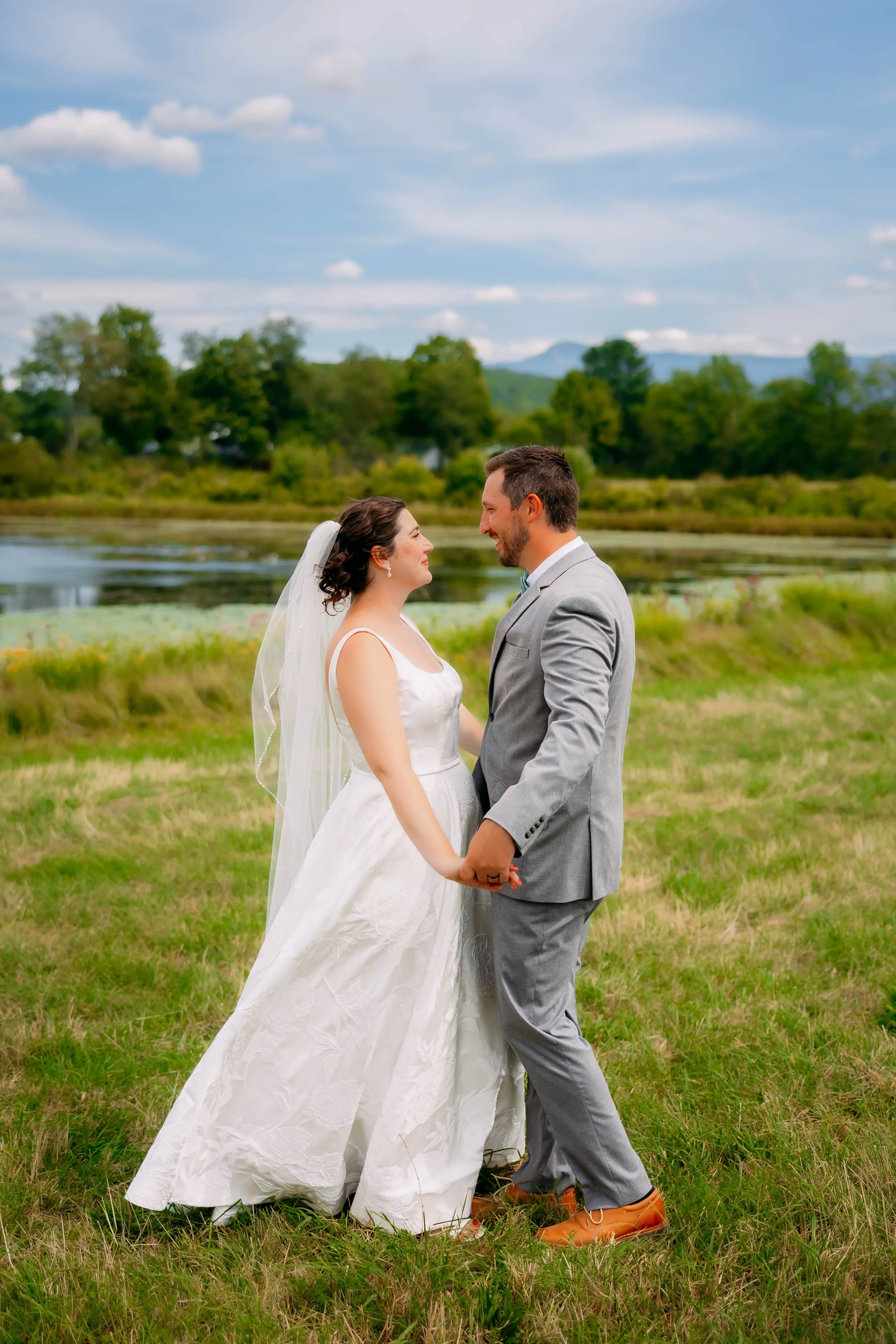 A newlywed couple holding hands and gazing at each other outdoors near a pond, with trees and mountains in the background on a partly cloudy day.