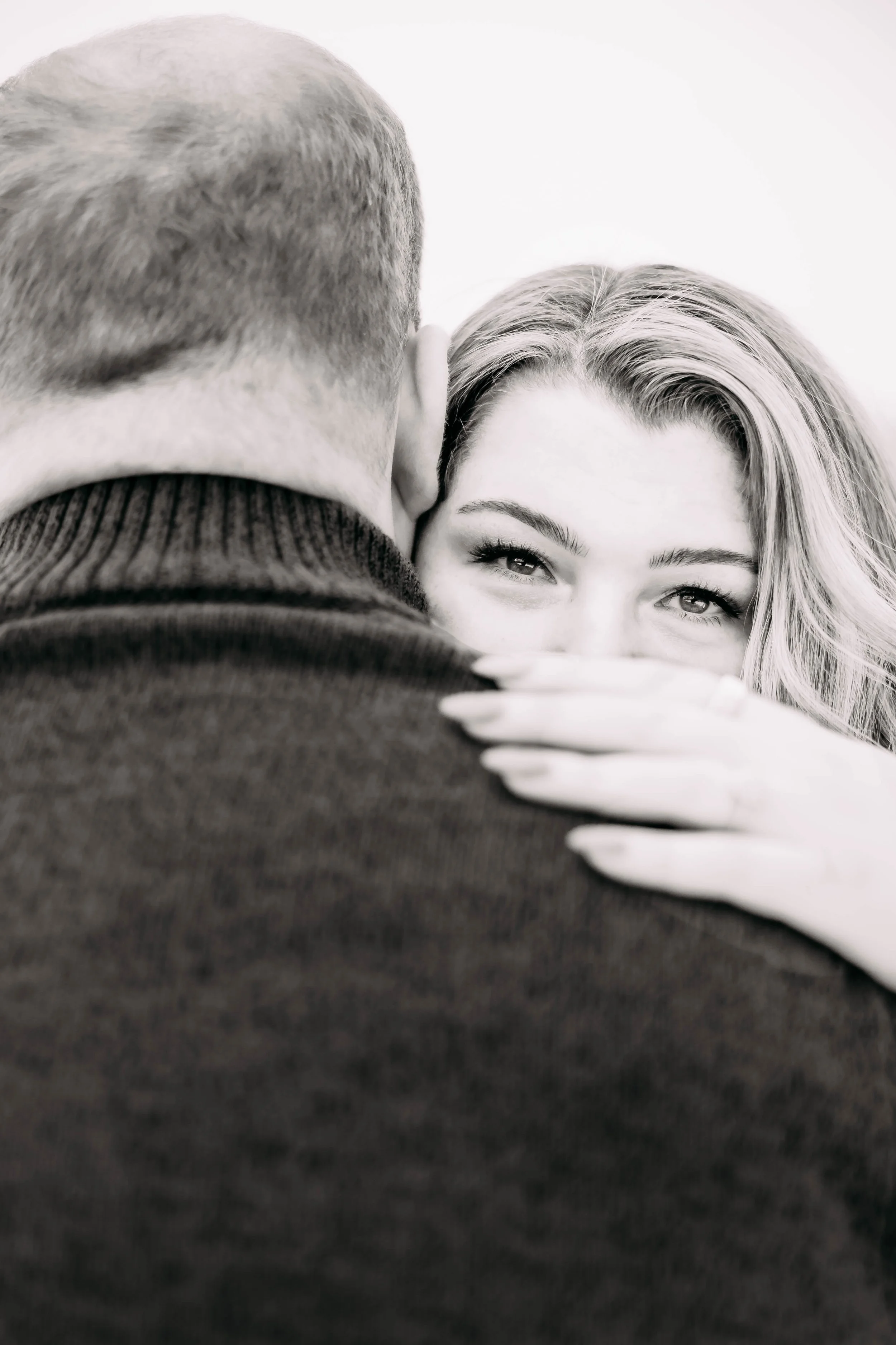 A woman embraces a man, her hand on his shoulder, with only the back of the man's head visible. The woman's eyes are smiling and she is partially hiding her face behind the man's shoulder.