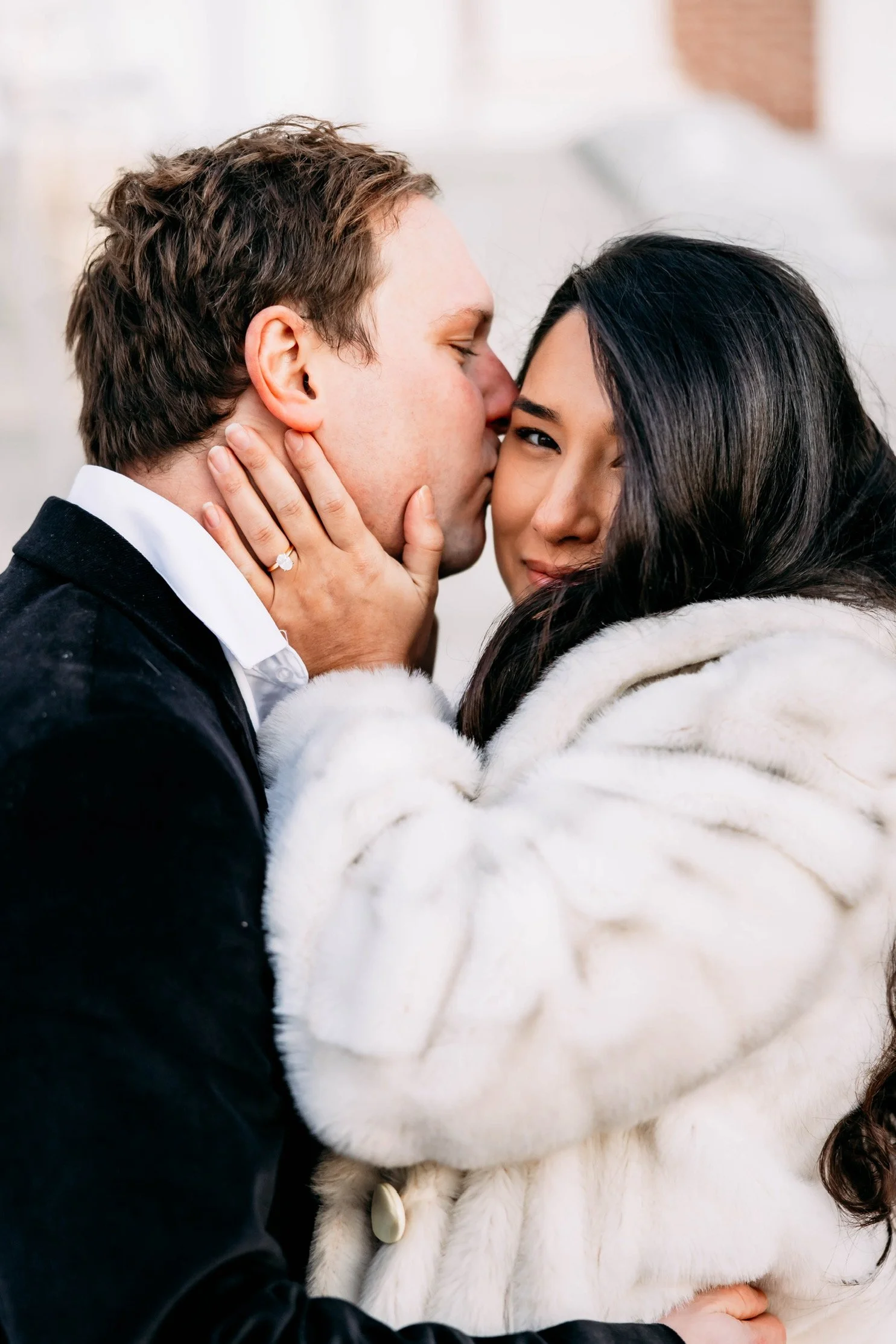 A man kissing a woman on the cheek while she smiles and holds his face, she is wearing a white fur coat and he is wearing a black jacket, in a close embrace.