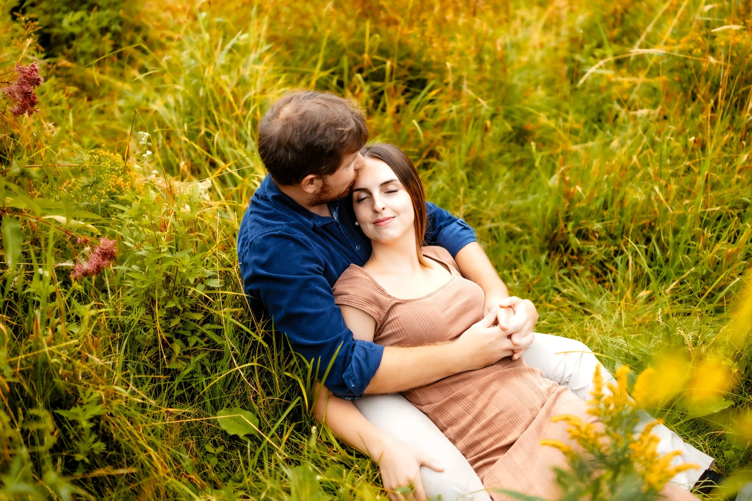 A couple sitting in a field of tall grass and wildflowers, with the man kissing the woman on the forehead. The woman is smiling with her eyes closed, wearing a brown dress, and the man is in a blue shirt.